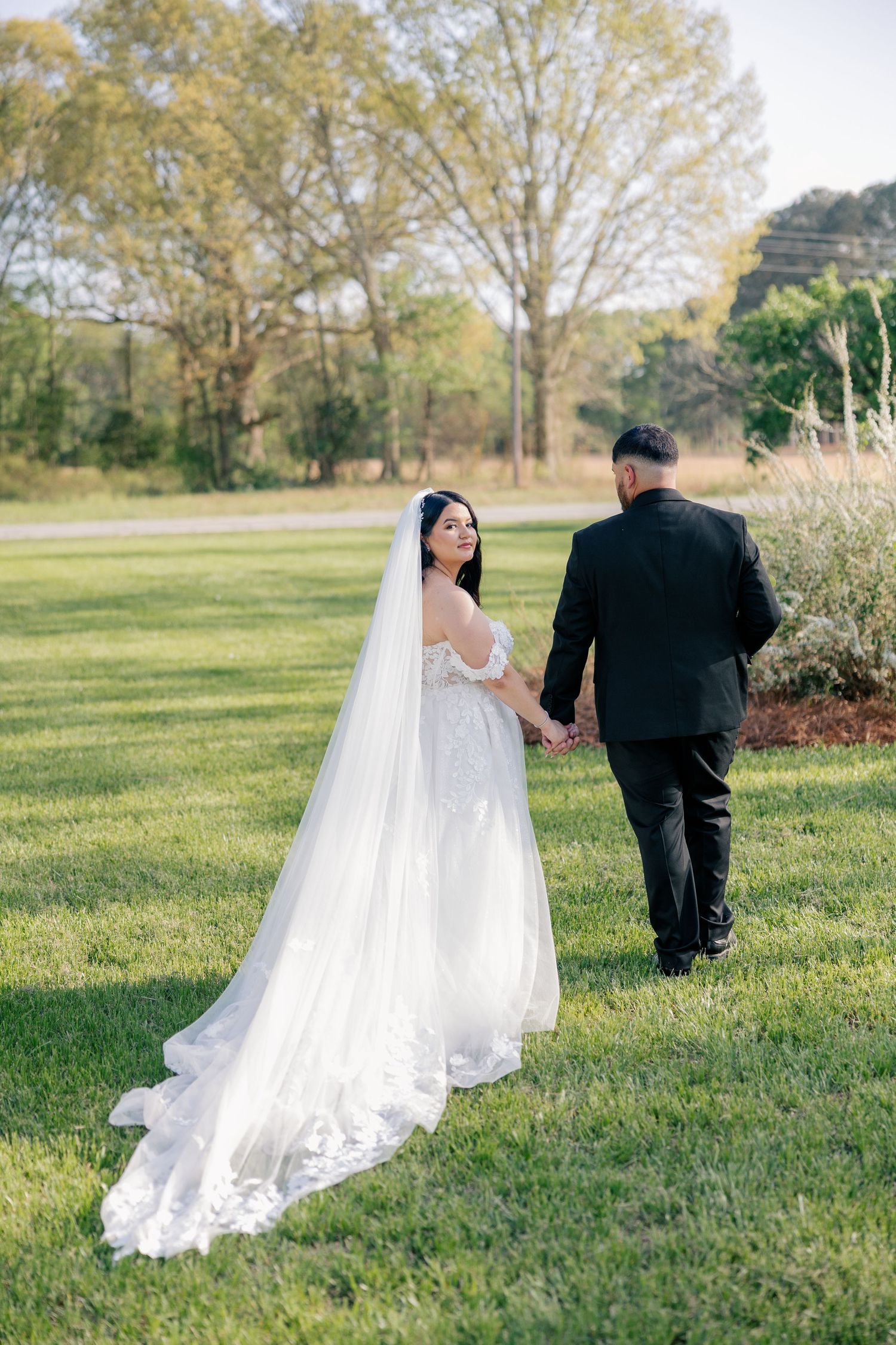 A couple in wedding attire walk through a grassy field as the bride's flowing white veil trails behind them.