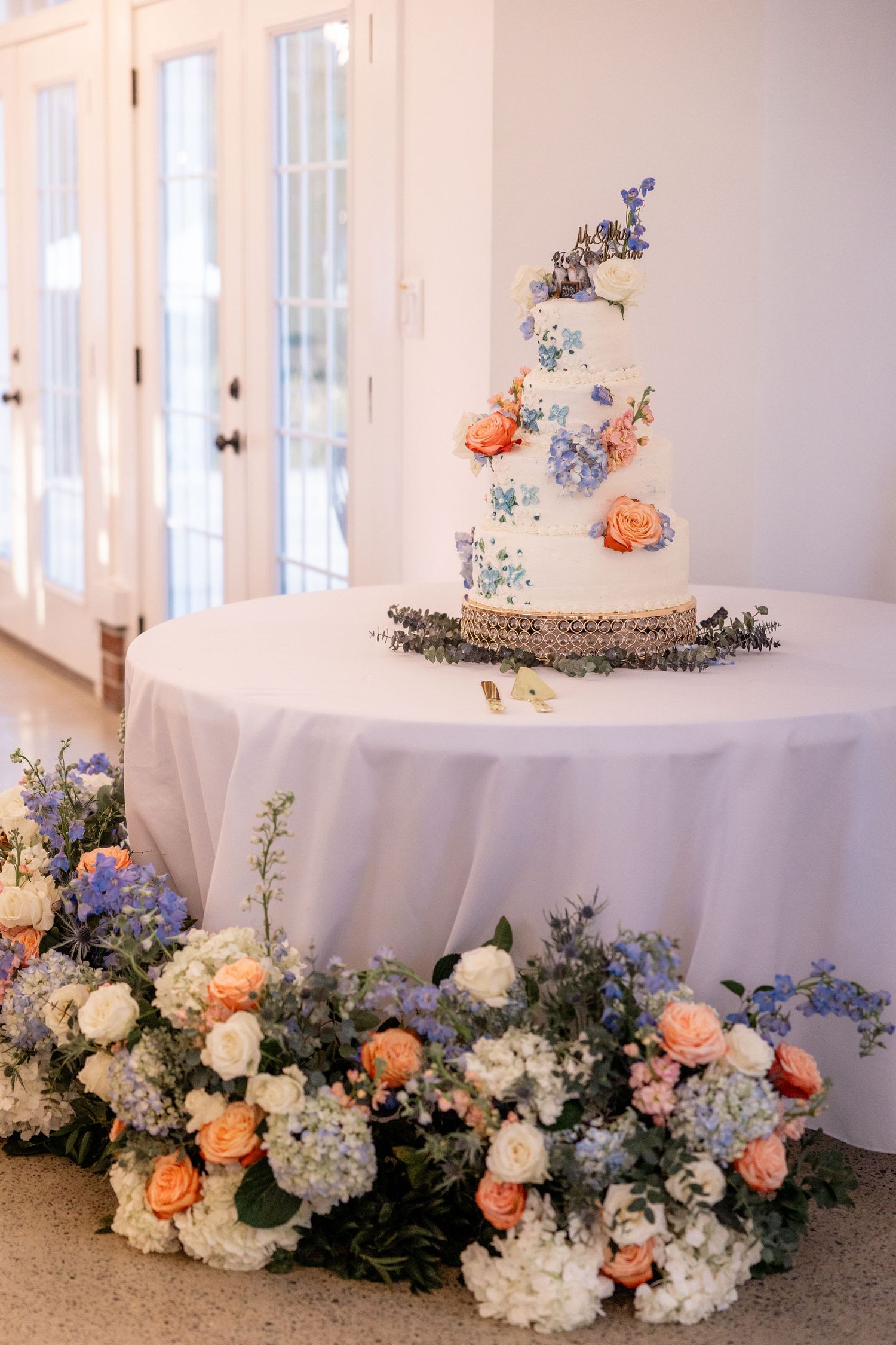Wedding cake table decorated with elegant floral arrangements in peach, blue and white blooms.