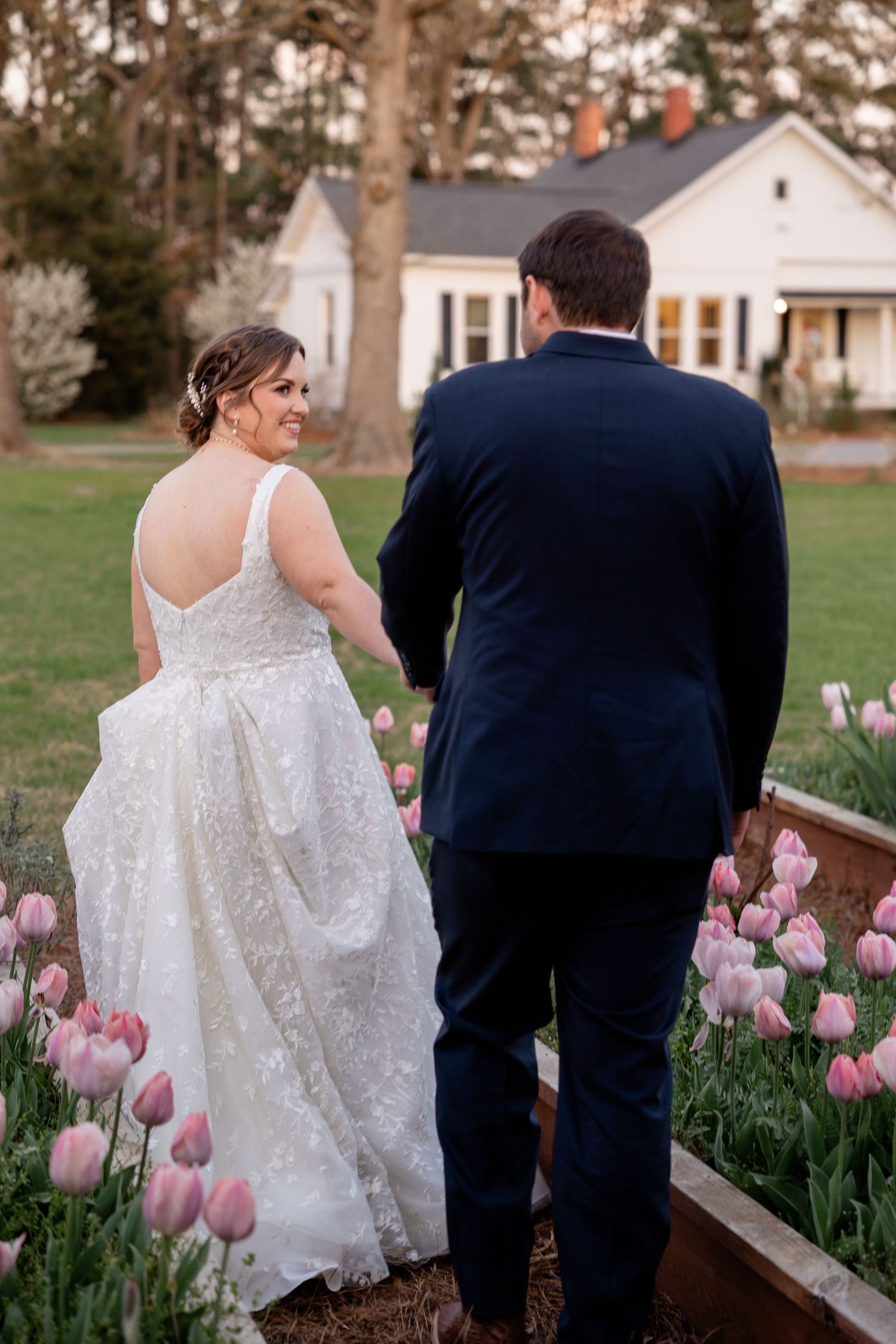 Wedding scene with a white dress and navy suit walking near a garden path lined with pink tulips.