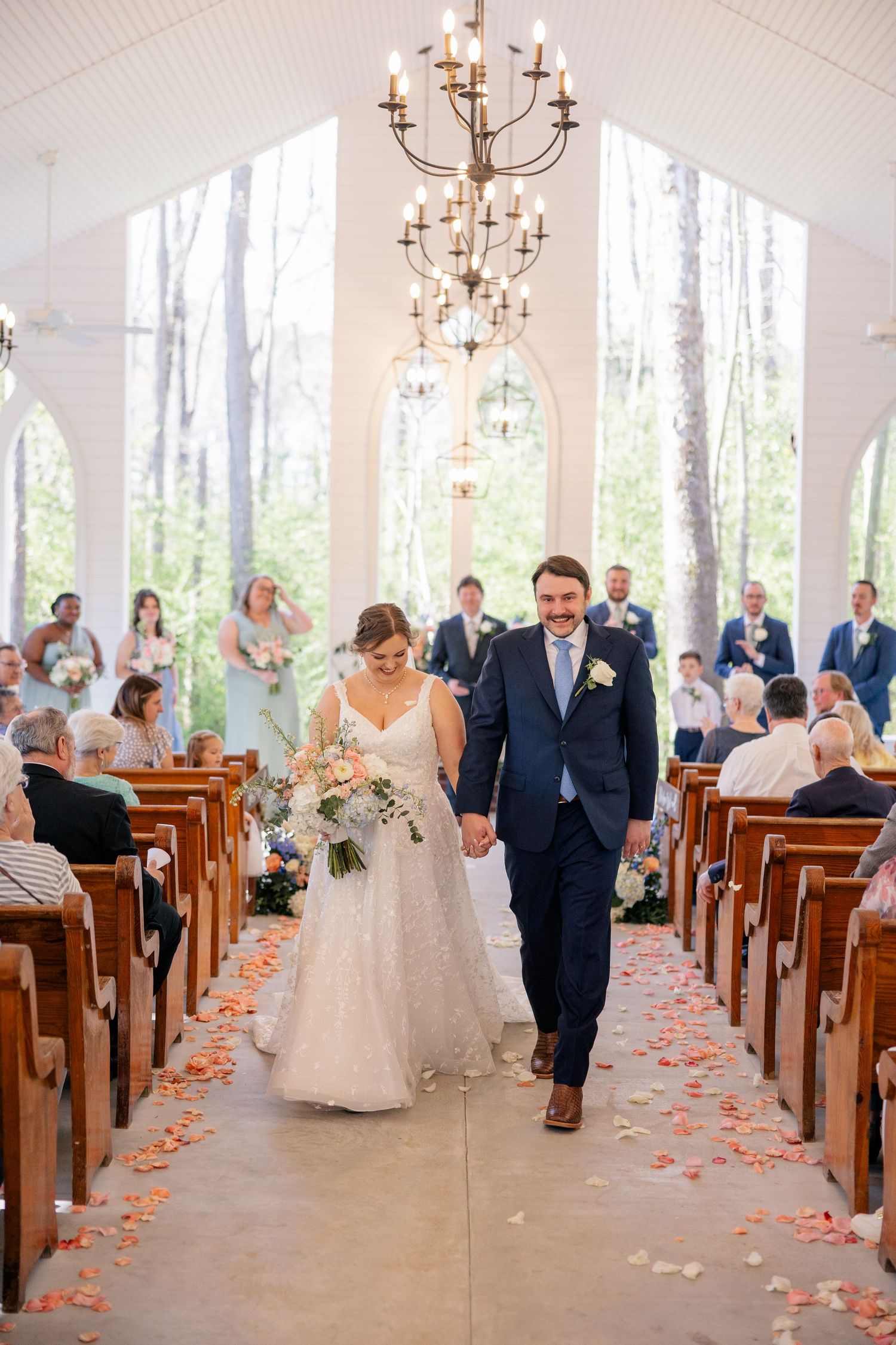 Wedding couple walks down aisle of chapel with wooden pews, chandeliers, and flower petals.