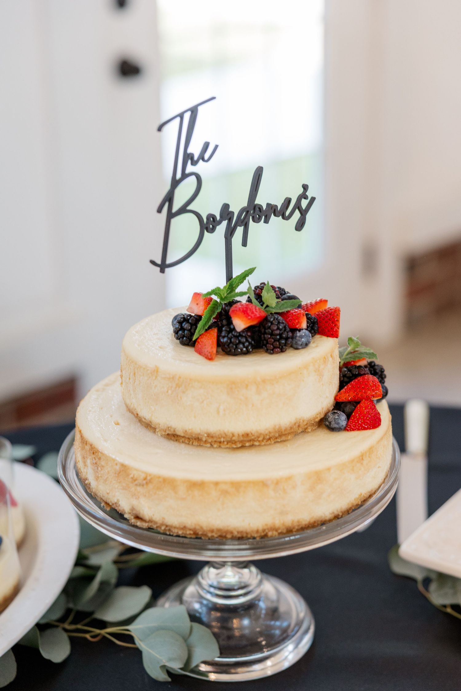 Two-tiered white cheesecake topped with fresh berries and a decorative cake topper on a glass stand.