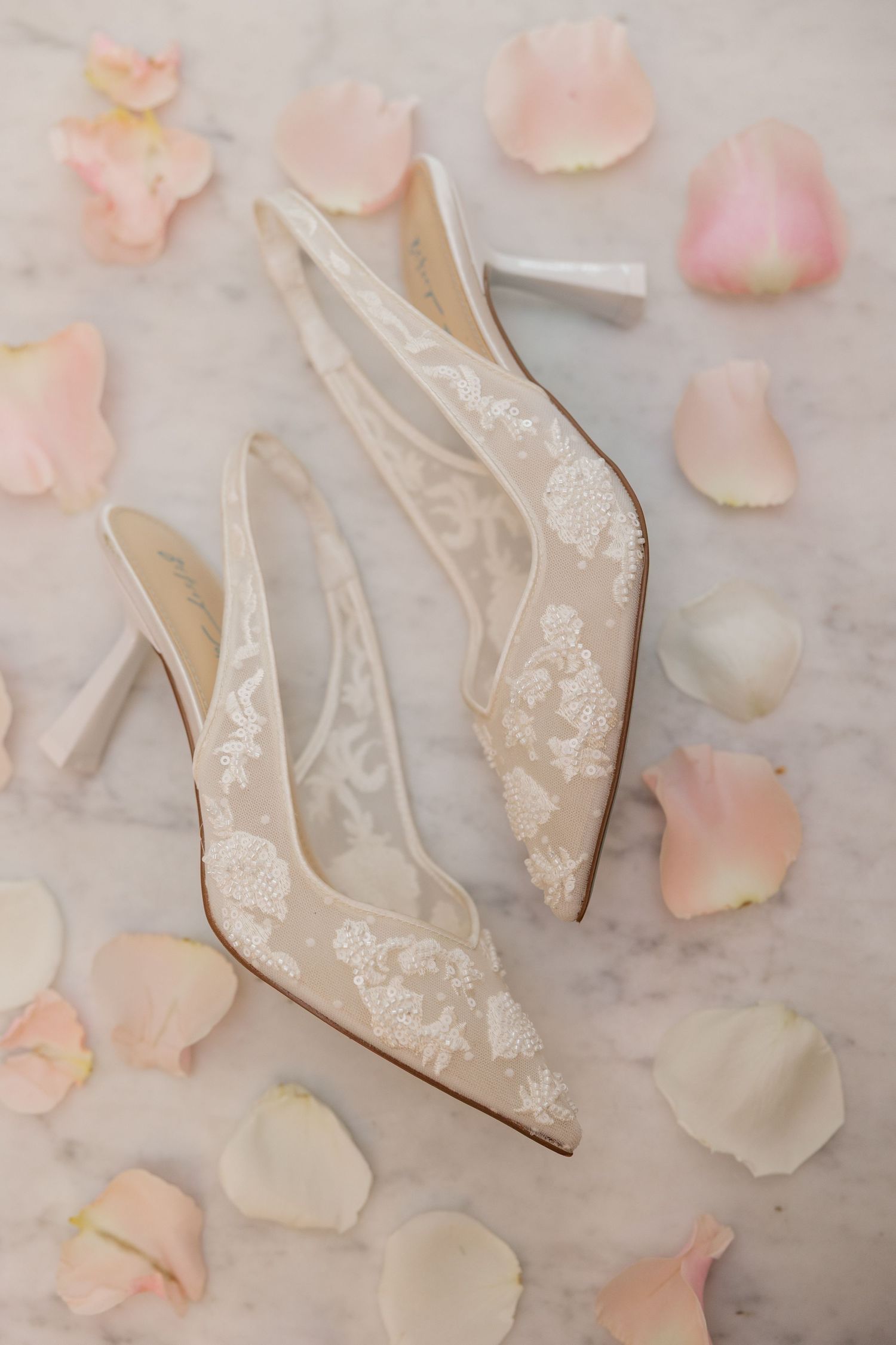 Elegant white lace wedding shoes arranged with scattered pink rose petals on a marble surface.