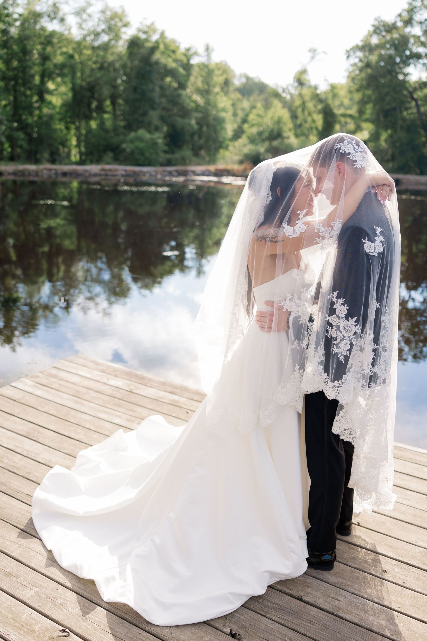 Wedding couple poses on wooden dock over reflective lake with flowing white veil in gentle breeze.