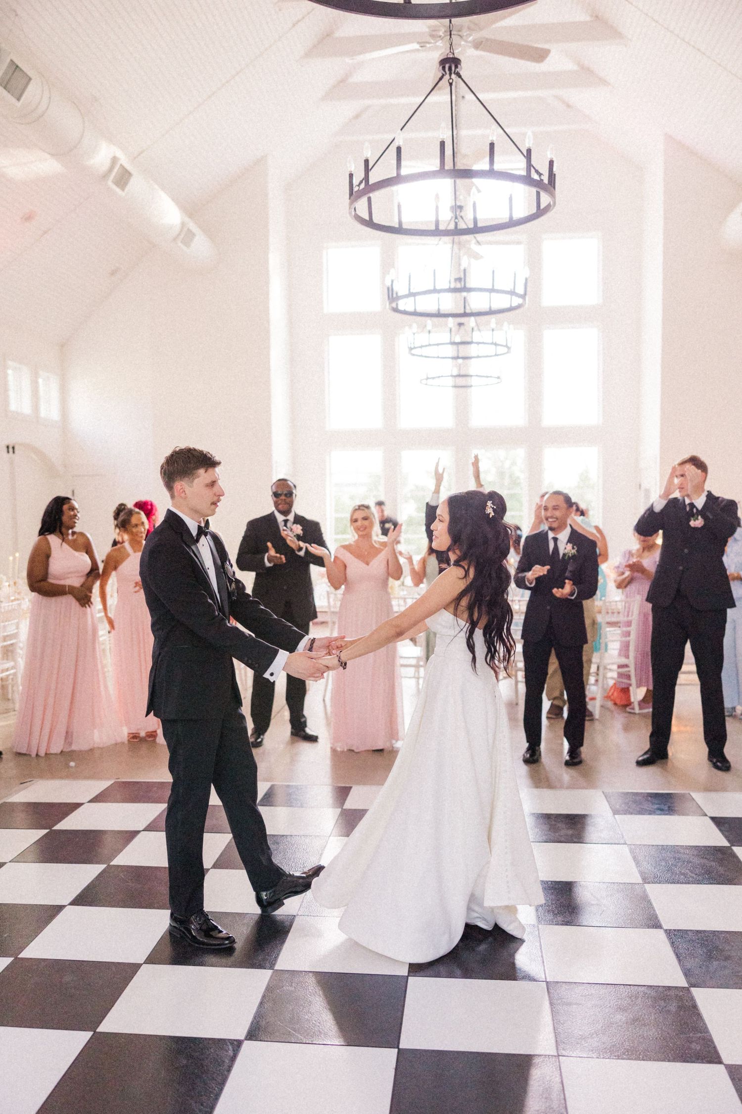 Wedding party dancing together on a black and white checkerboard dance floor in an elegant white venue.
