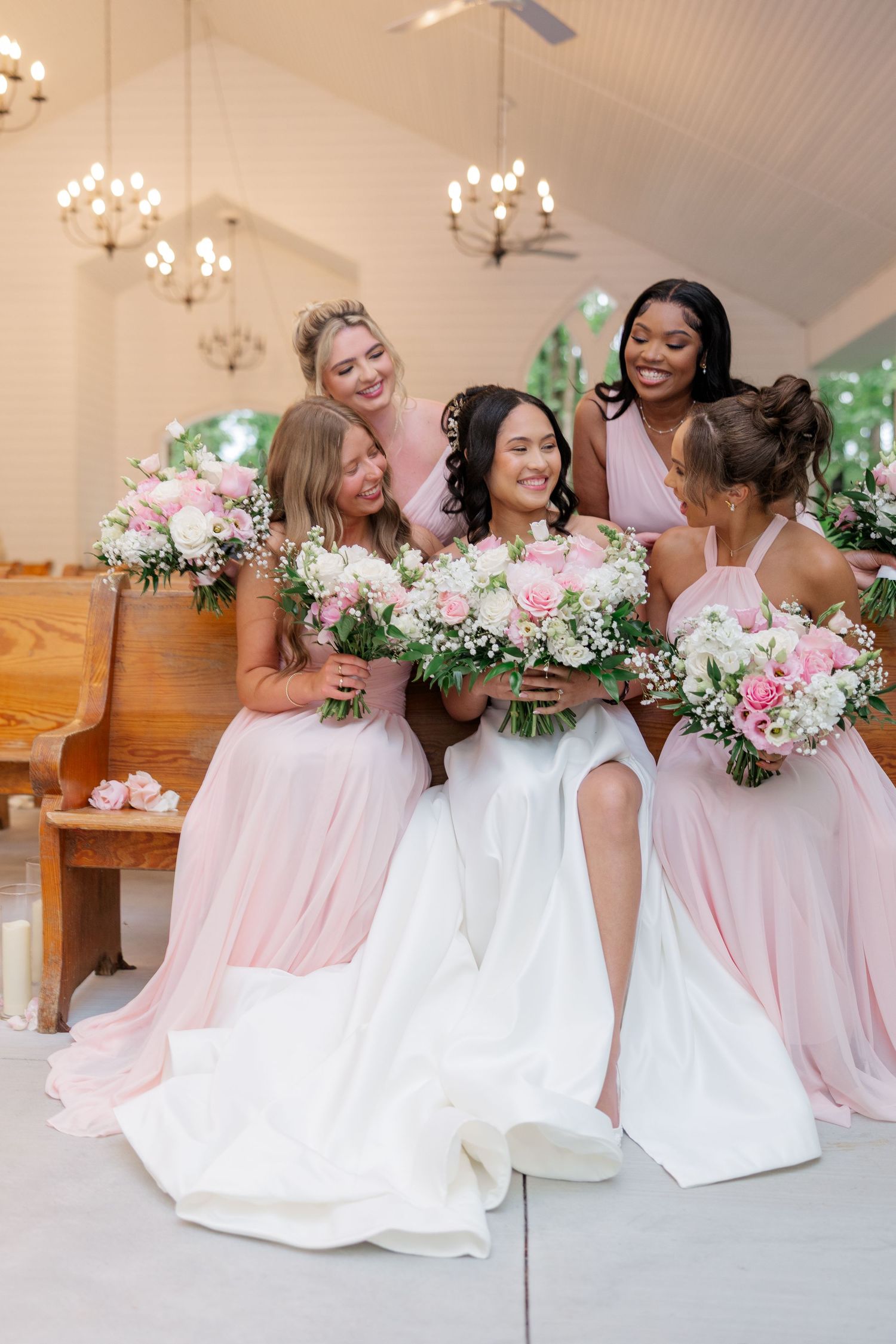 Bridesmaids in blush pink dresses holding pink and green bouquets share joyful laughter during wedding photos.