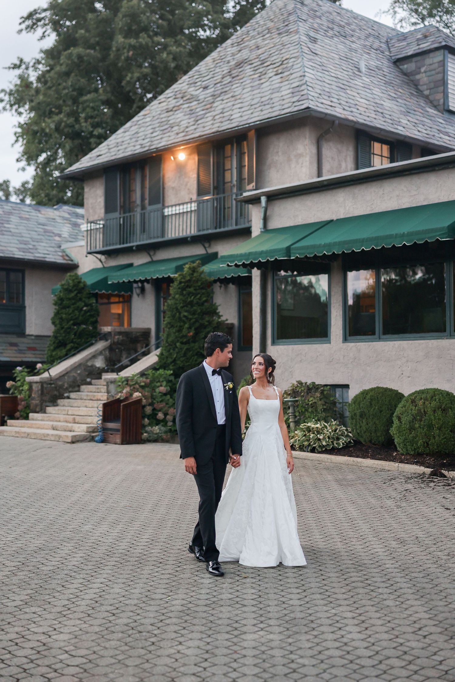Bride and groom hold hands walking on cobblestone path before elegant stone manor with green awnings.
