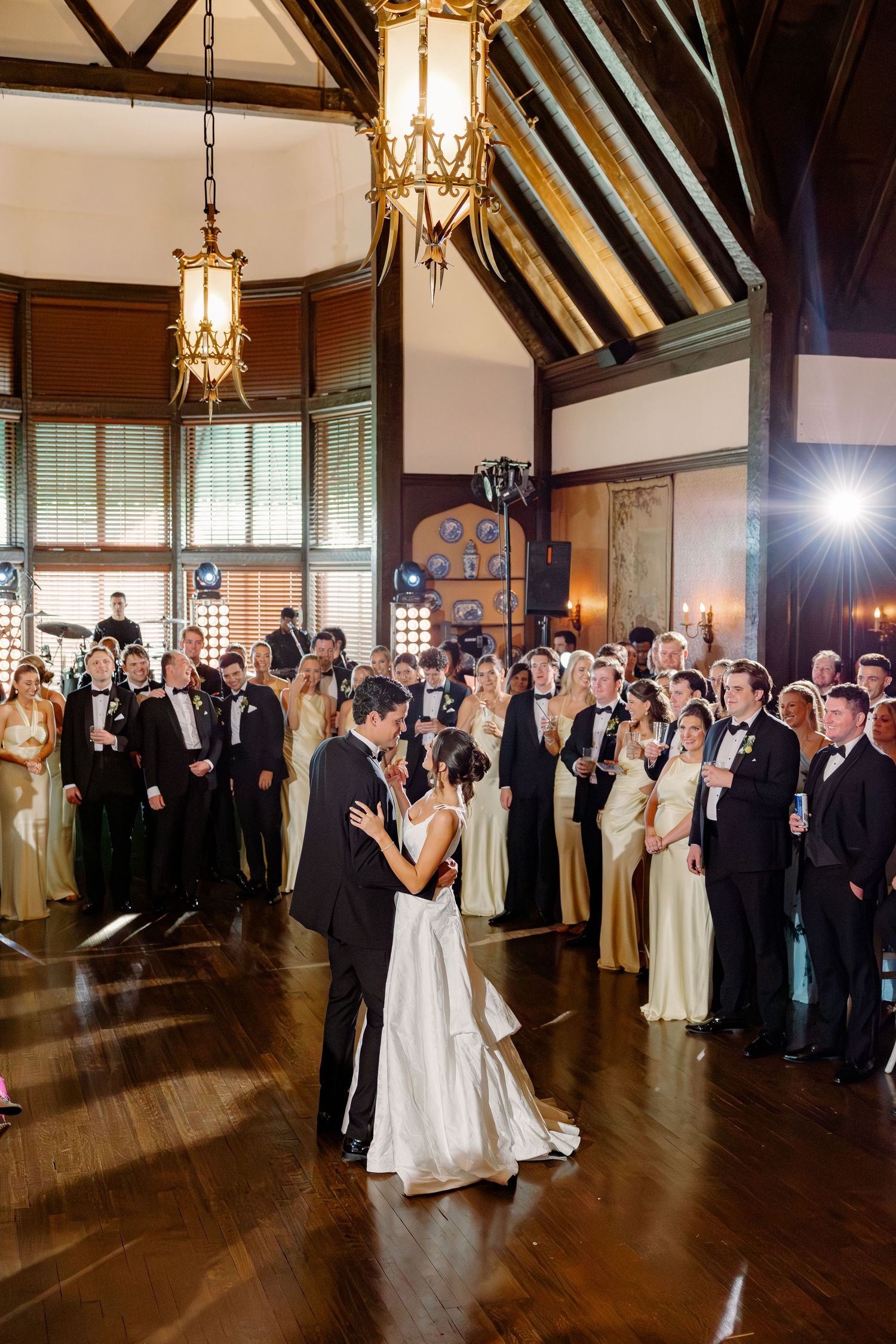Bride and groom share first dance surrounded by guests in formal attire in a grand ballroom.