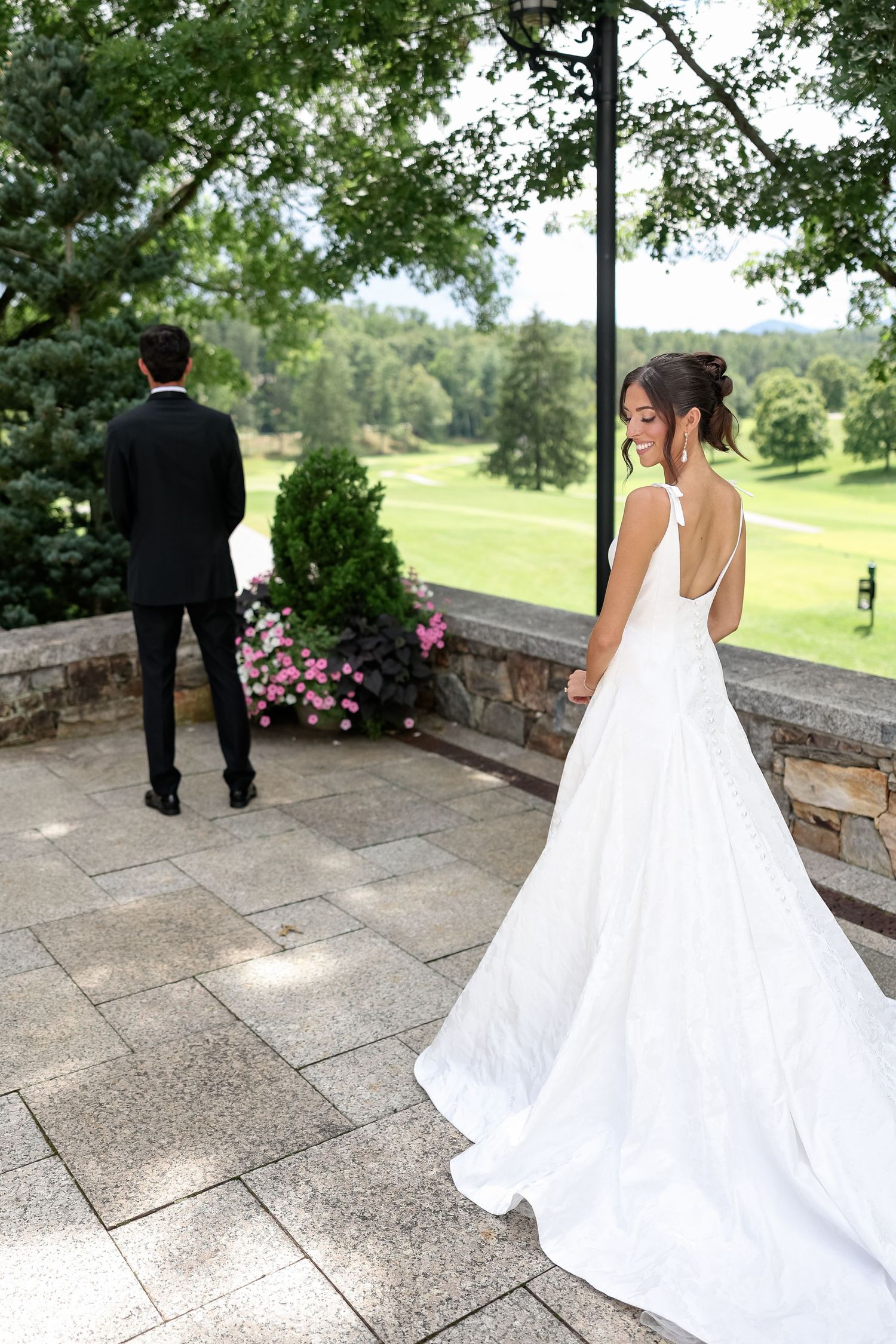Bride in white gown smiles over shoulder as groom stands at stone terrace overlooking lush green golf course.