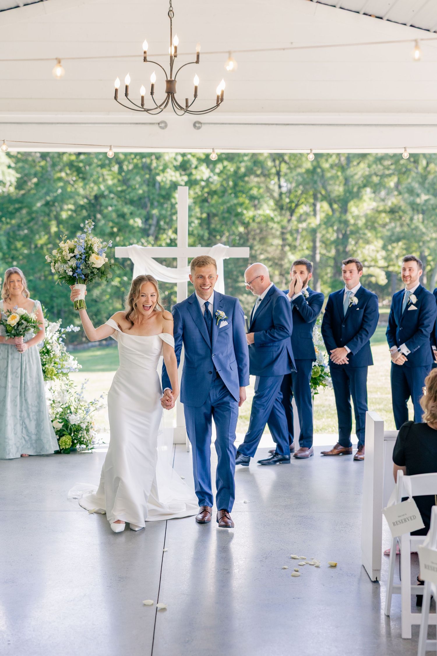 Newlyweds walk down aisle in elegant white tent venue with cross backdrop and cheering wedding party.
