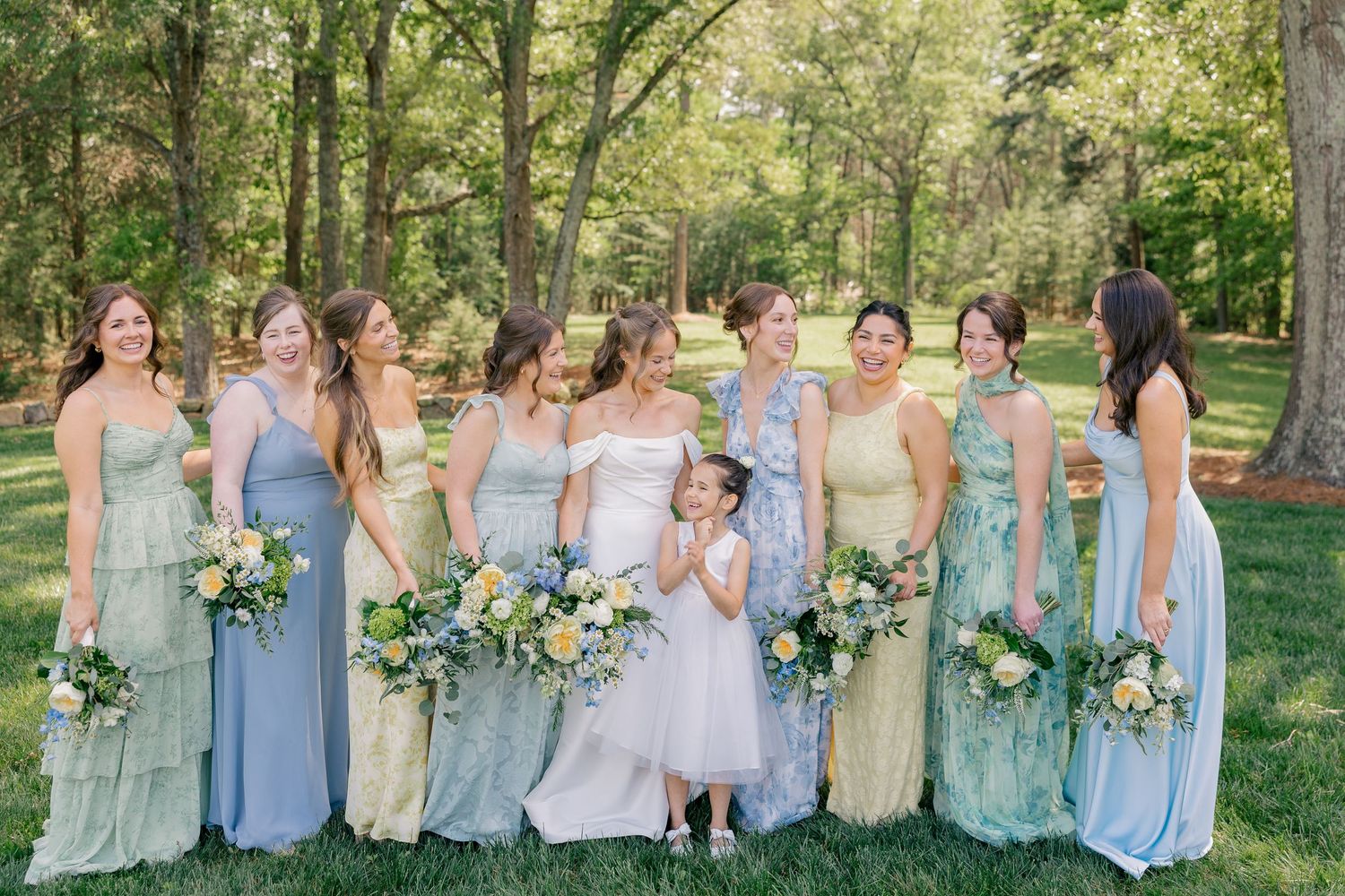 Bridesmaids in mismatched blue and green gowns hold bouquets with bride and flower girl outdoors.