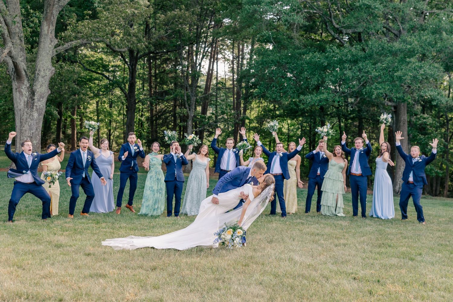 Wedding party celebrates with dip kiss pose on green lawn surrounded by tall trees at outdoor reception.