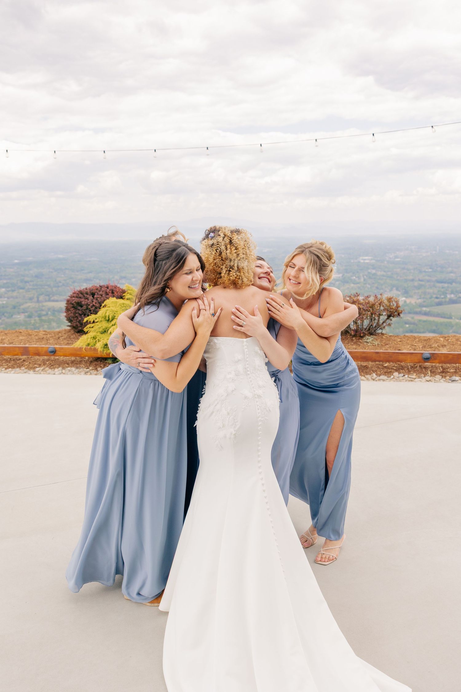 Bride in white gown embraced by bridesmaids in dusty blue dresses at scenic outdoor venue.