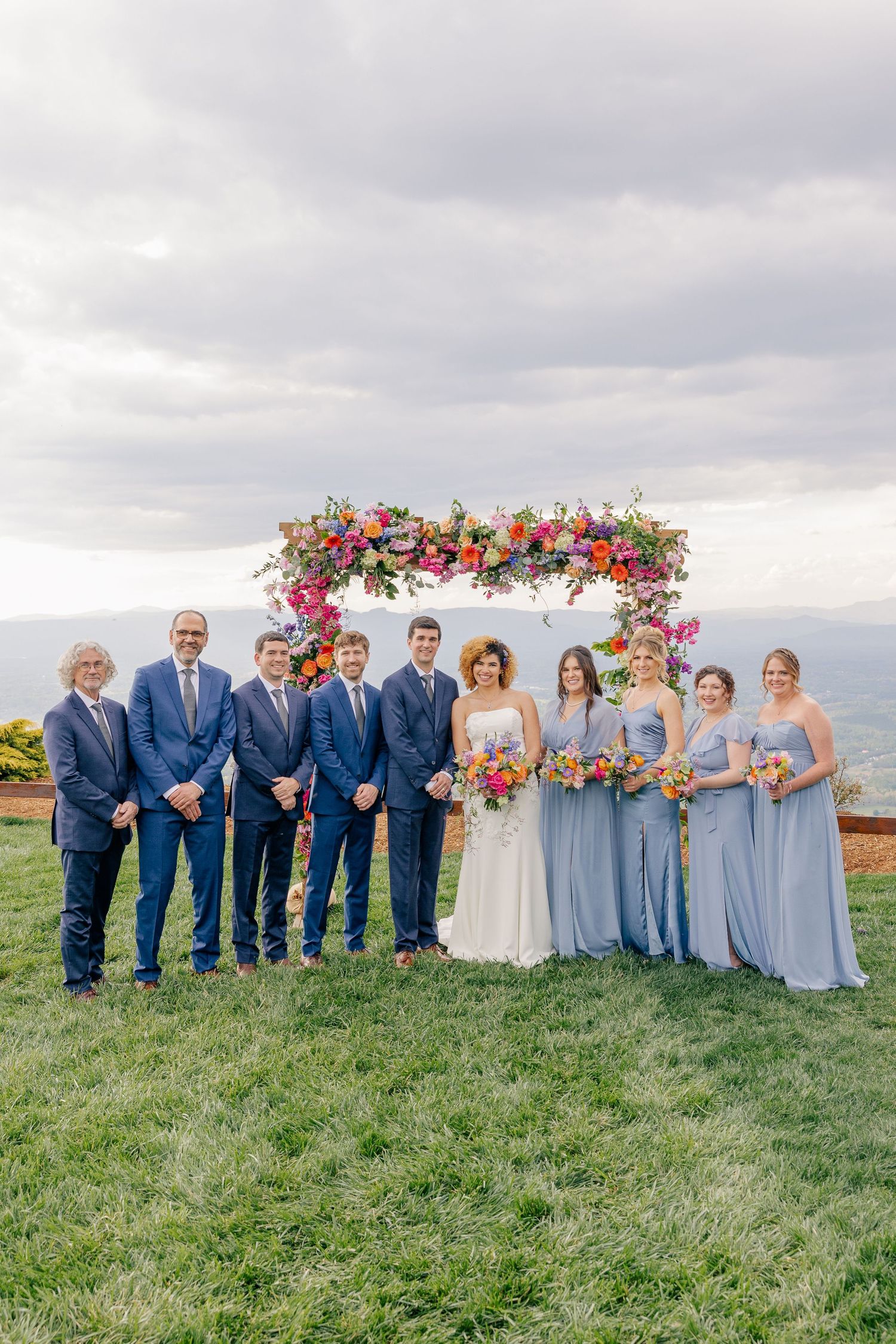 Wedding party poses in front of colorful floral arch with bridesmaids in blue gowns and groomsmen in navy suits.
