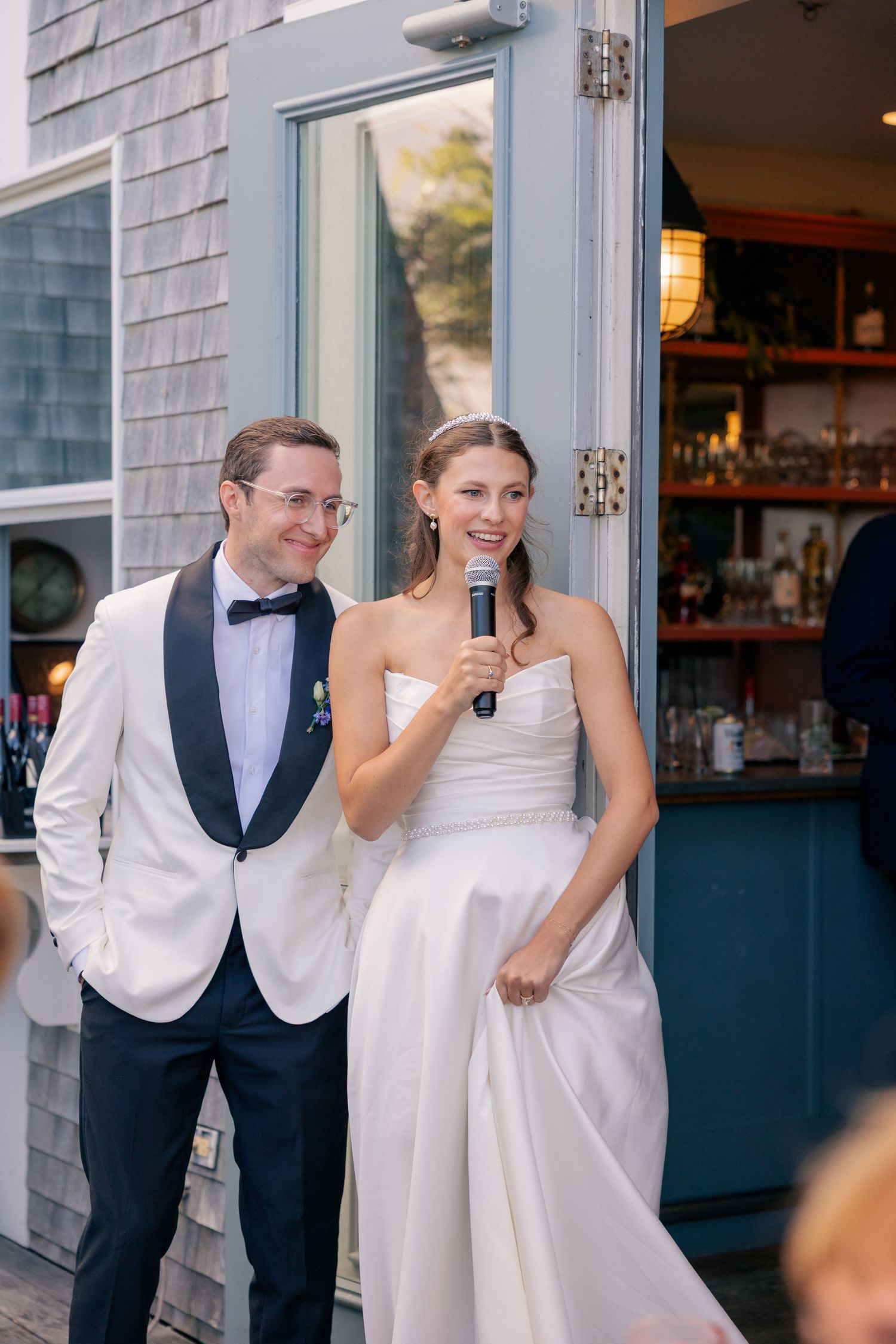 Bride speaks into microphone while groom smiles beside her at outdoor wedding reception venue.