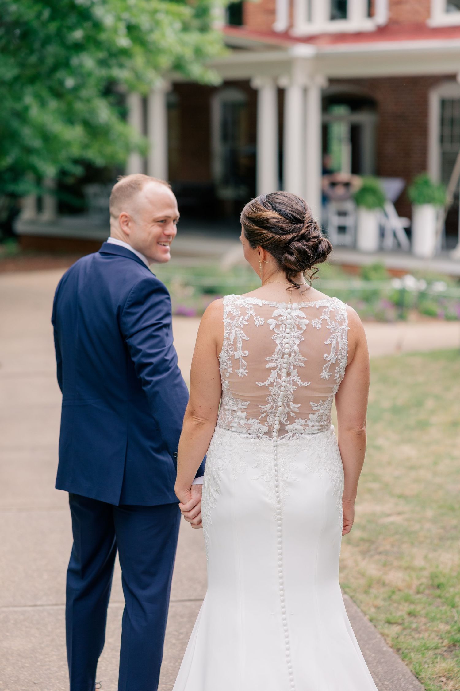 Bride and groom holding hands walking toward a brick mansion, showcasing the lace back wedding gown and navy suit.