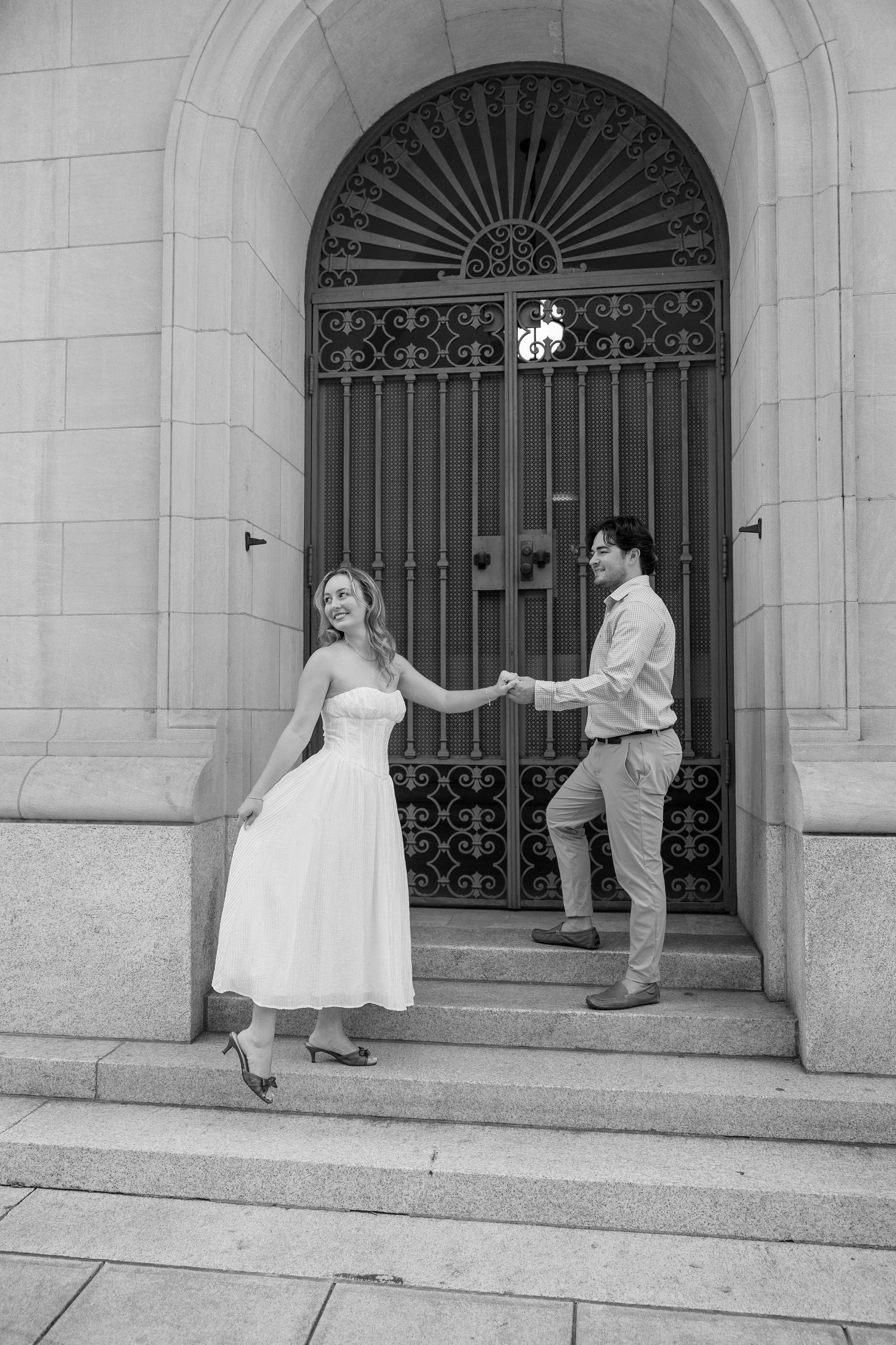 Black and white photo of a couple holding hands on stone steps before an ornate arched iron gate.