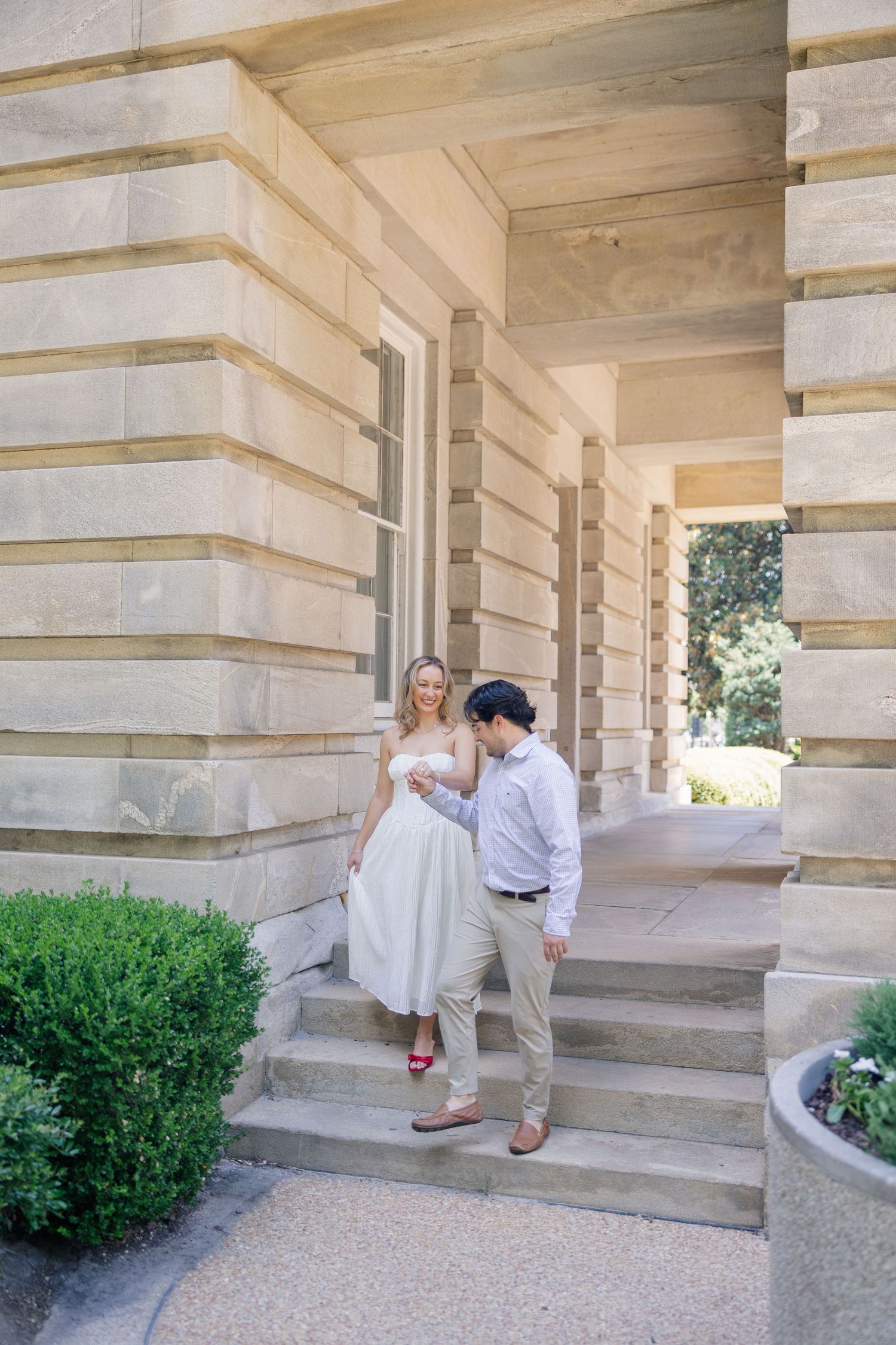 Engaged couple sharing a tender moment on stone steps of a classic sandstone building with lush greenery.