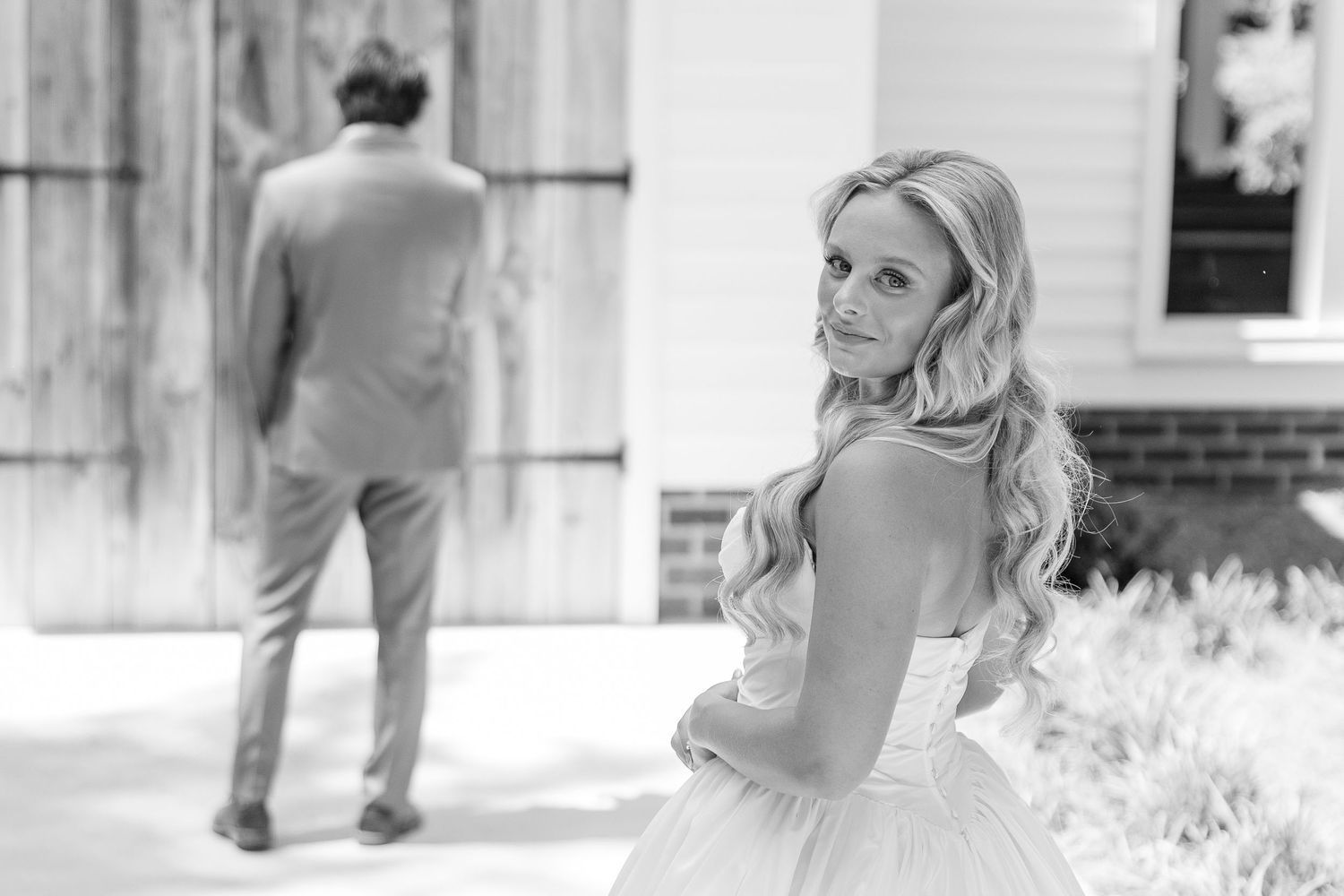 Black and white photo of smiling bride in strapless gown with groom standing ahead at barn venue.