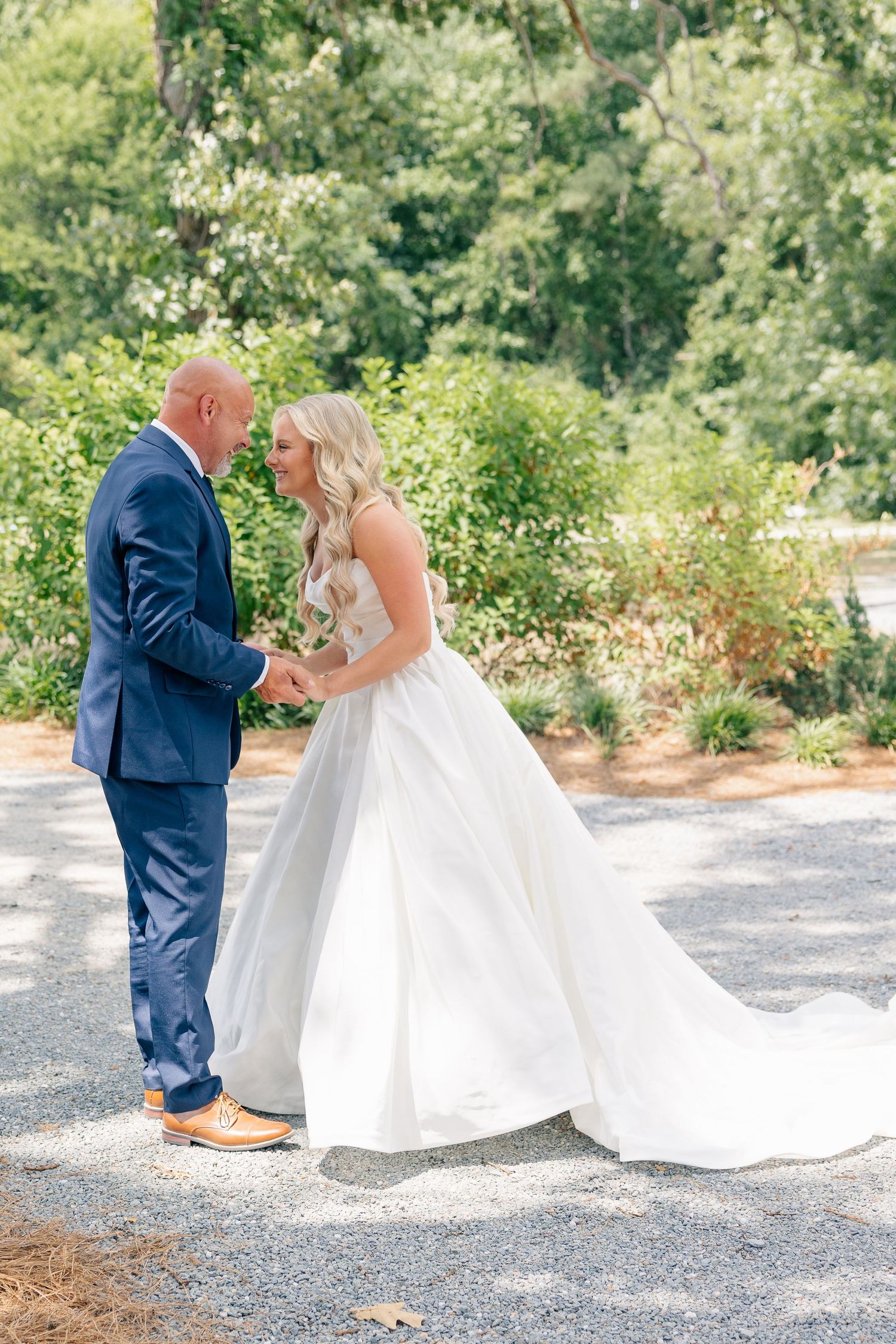 A bride in a white ballgown holds hands with a groom in a navy suit outdoors at a garden wedding venue.