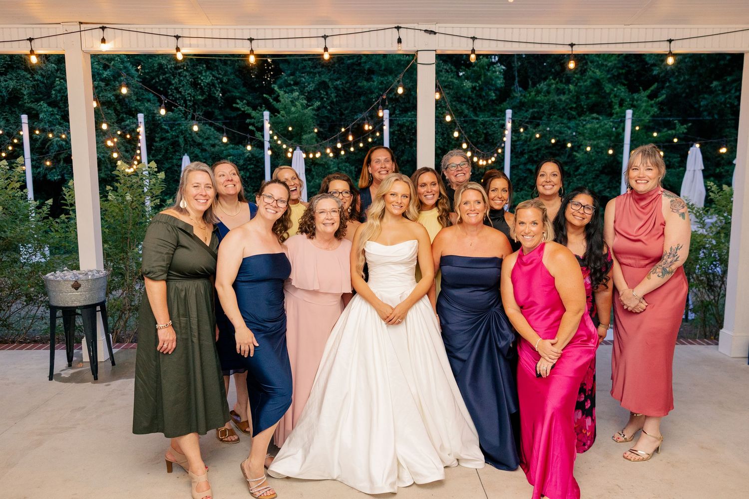 Bride in white gown posing with group of women in colorful dresses at evening reception.