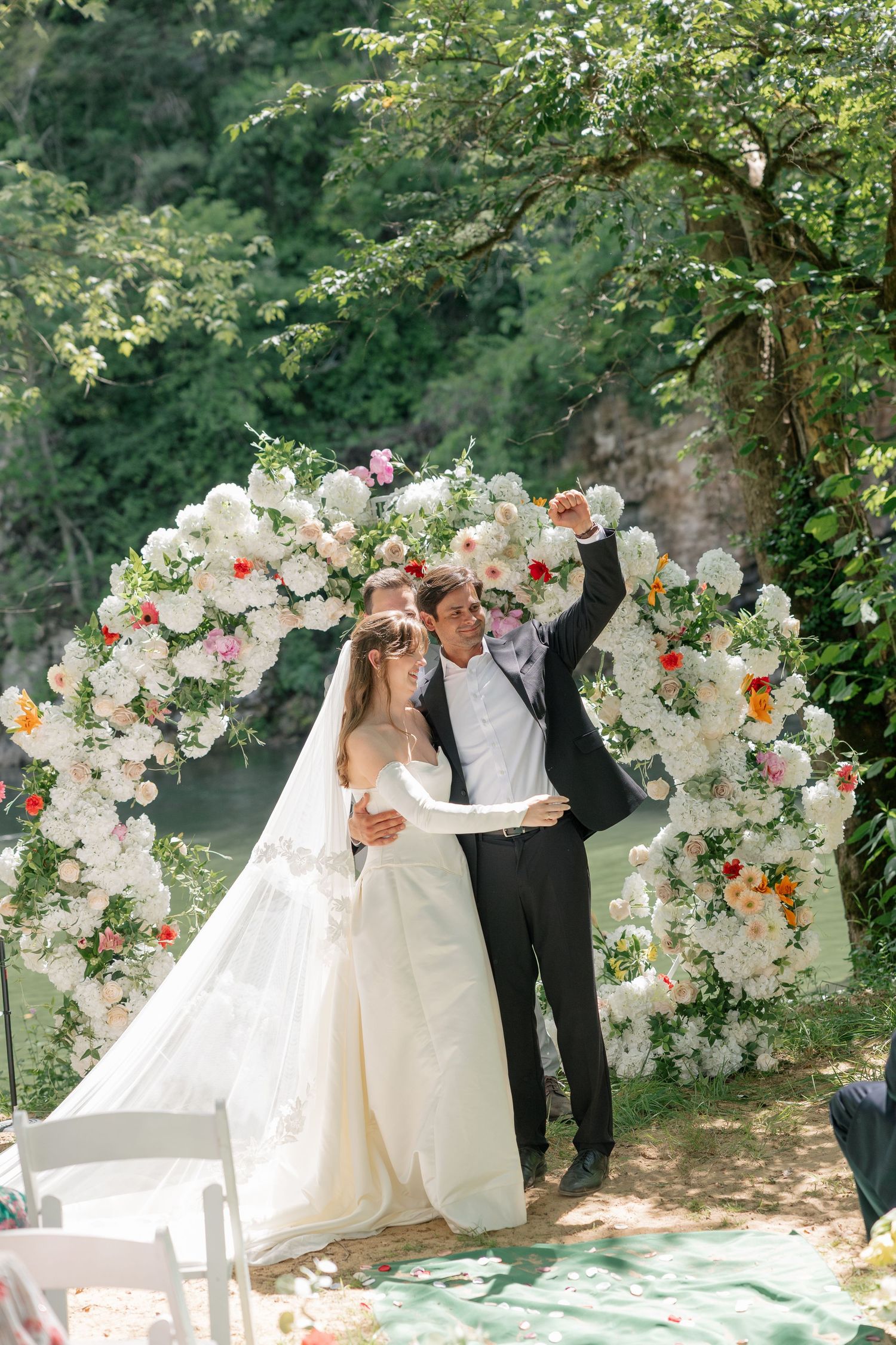 Joyful newlyweds celebrate with raised arms before a lush floral arch at their outdoor riverside ceremony.
