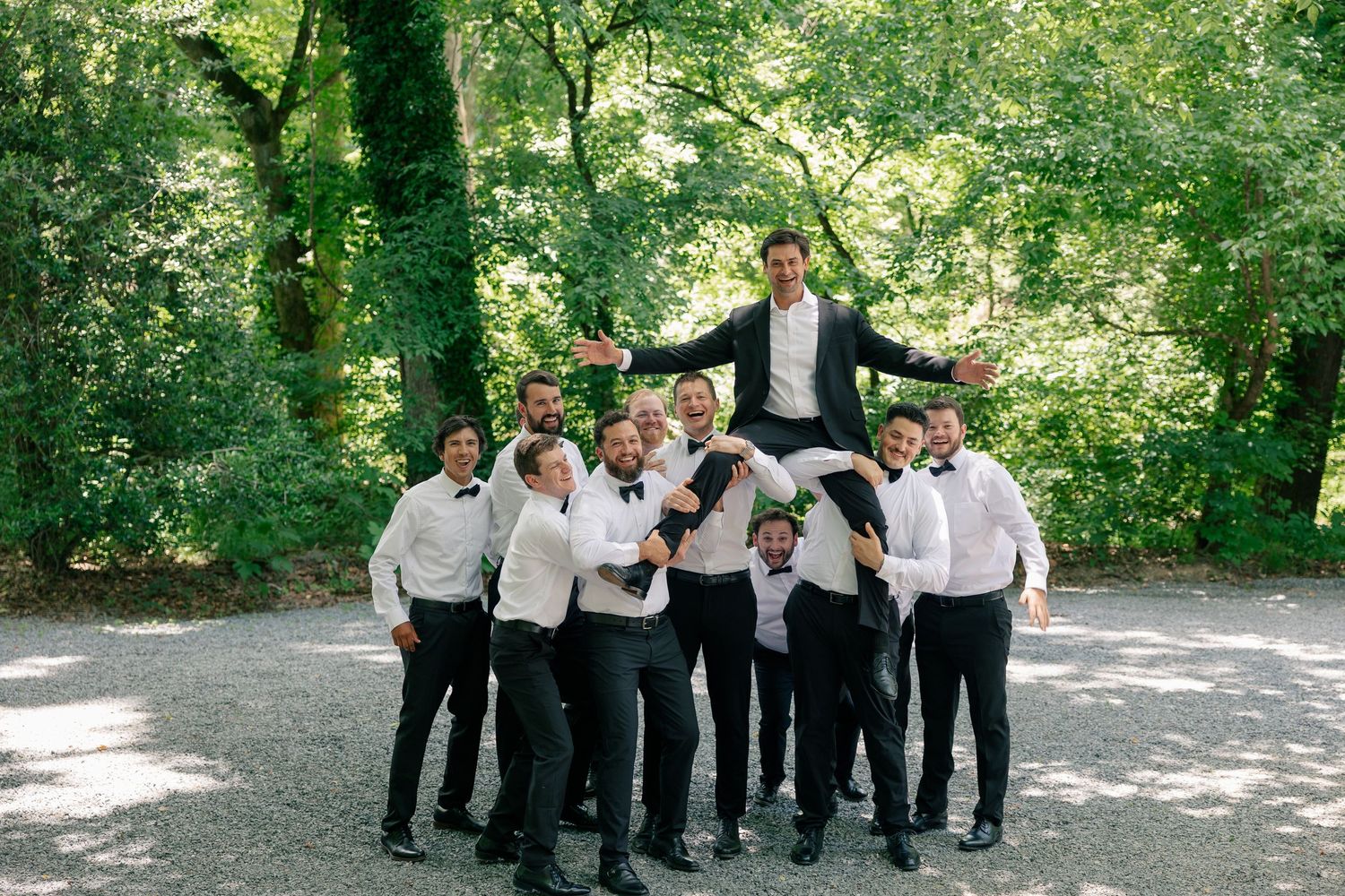 Groom lifted by groomsmen in white shirts and black suits outdoors, celebrating with joy in a lush green garden.