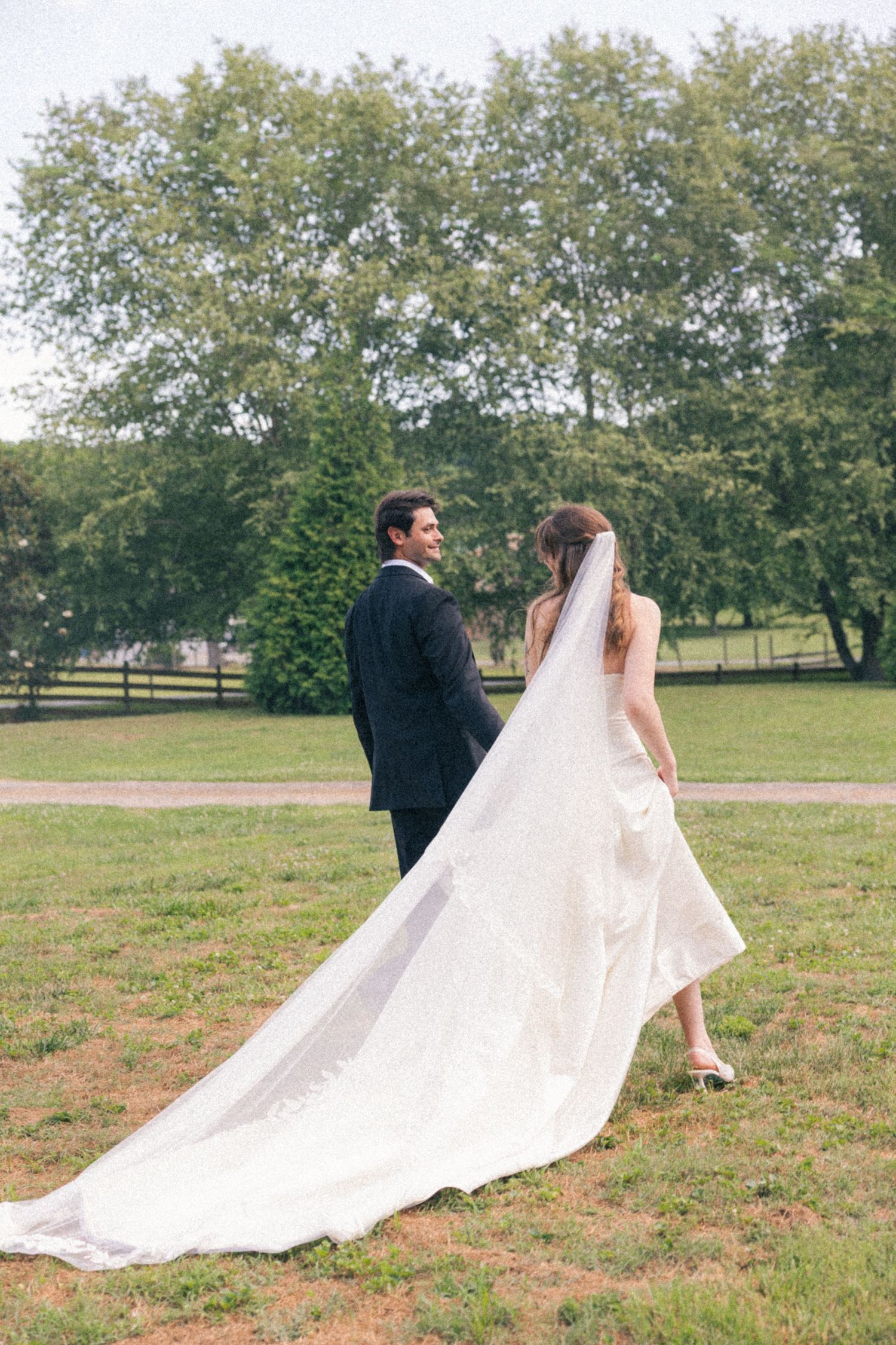Bride and groom walk hand in hand across a lush green lawn, her cathedral veil trailing behind.