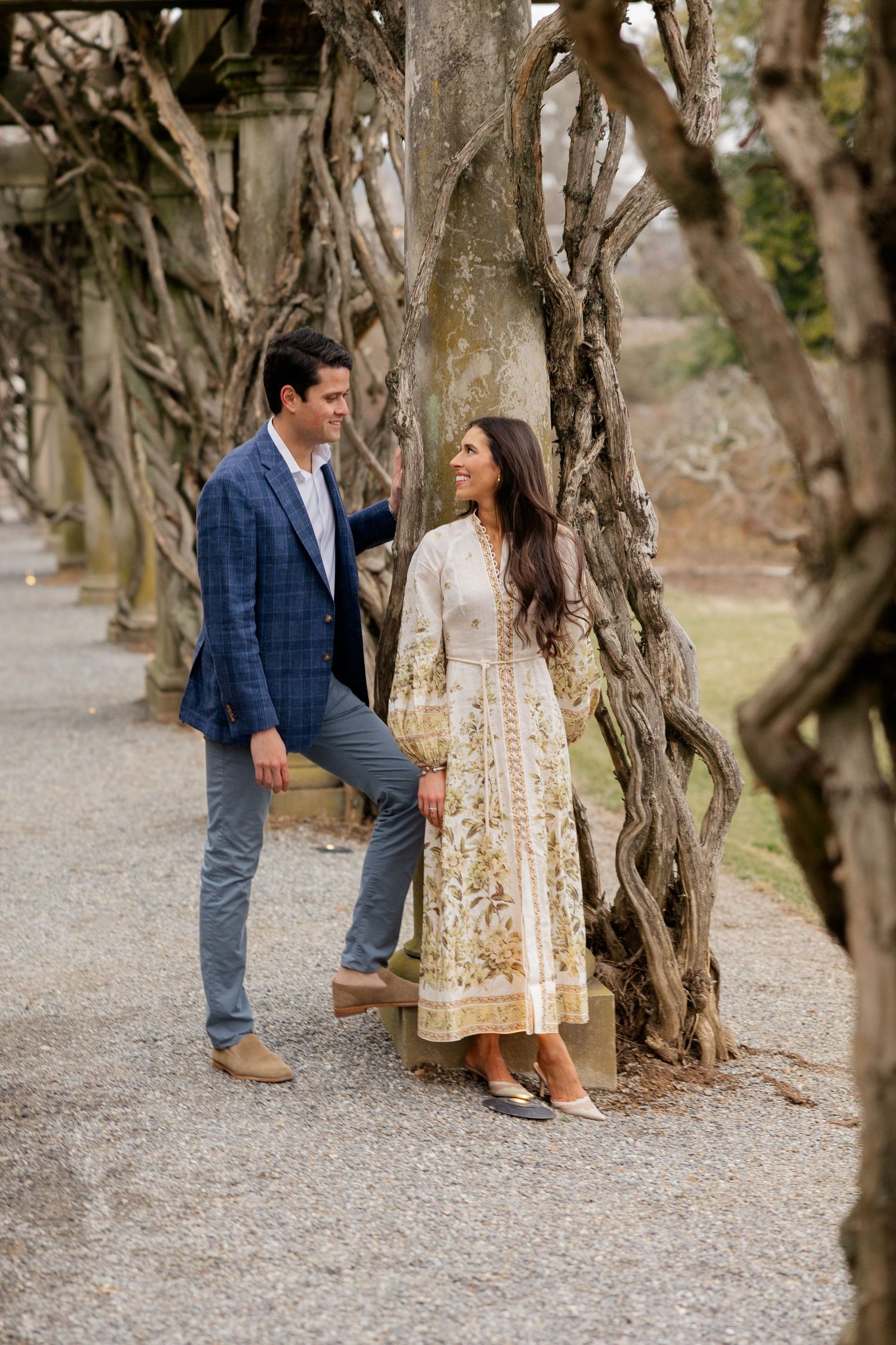A couple stands together on a path lined with twisted trees as she wears traditional South Asian dress and he wears a blazer.