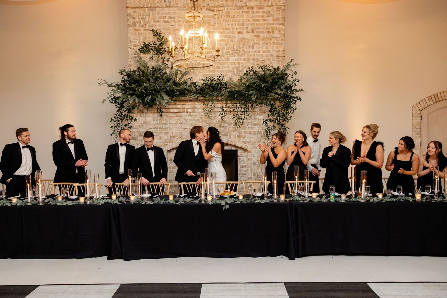 Wedding party seated at long table with elegant greenery and chandelier decor against brick wall.
