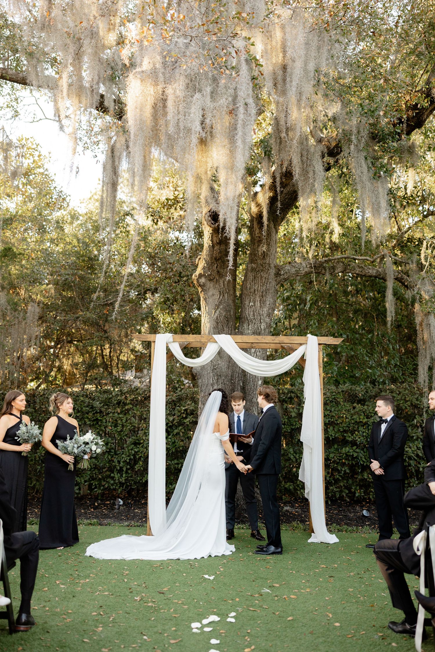 A romantic outdoor wedding ceremony under Spanish moss with black bridesmaid dresses and white floral arch.
