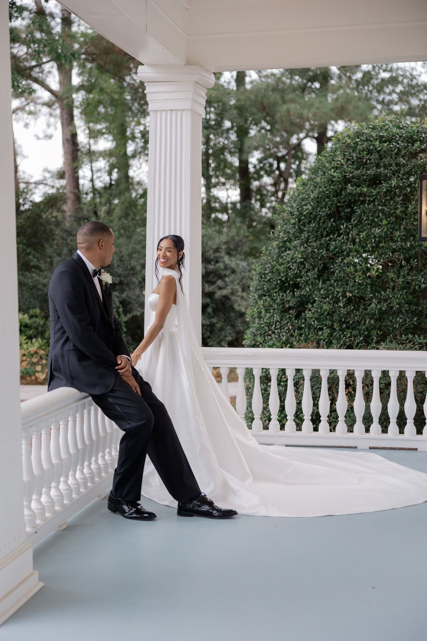 A bride and groom share a playful moment on a white columned porch during their wedding photoshoot. Cornealius Properties Wedding Photograph