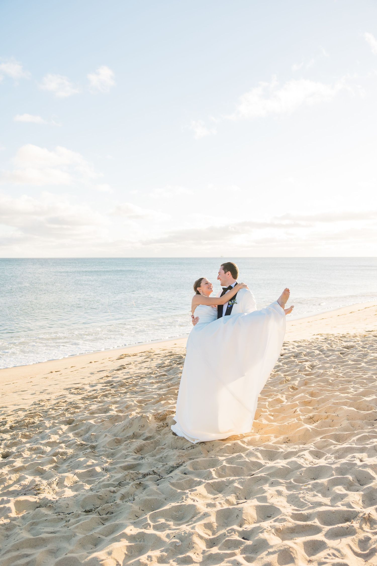 Groom carrying bride in white dress on sandy beach with ocean backdrop at sunset wedding.