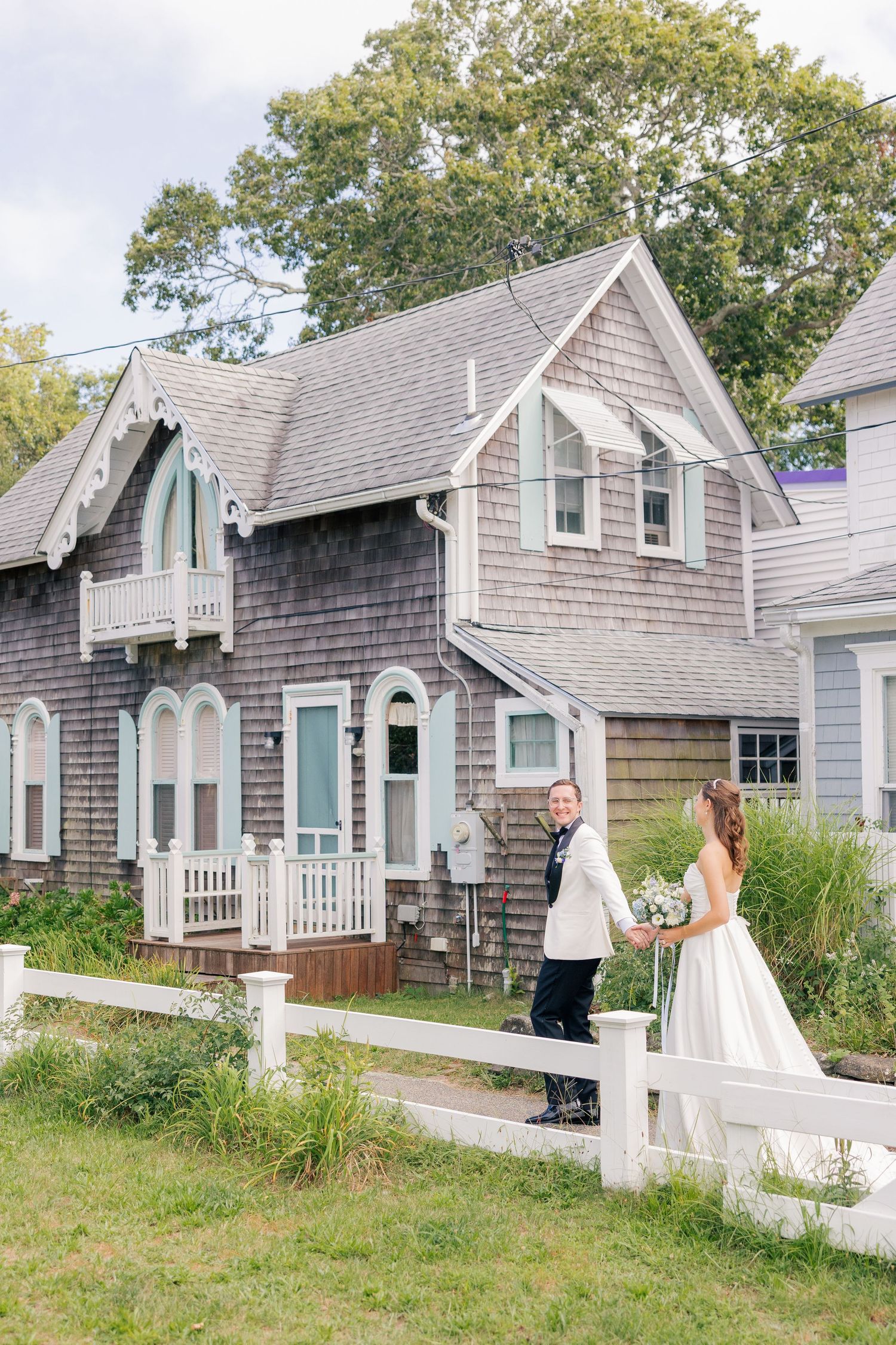 Bride and groom walking along white picket fence in front of charming shingle-style cottage.
