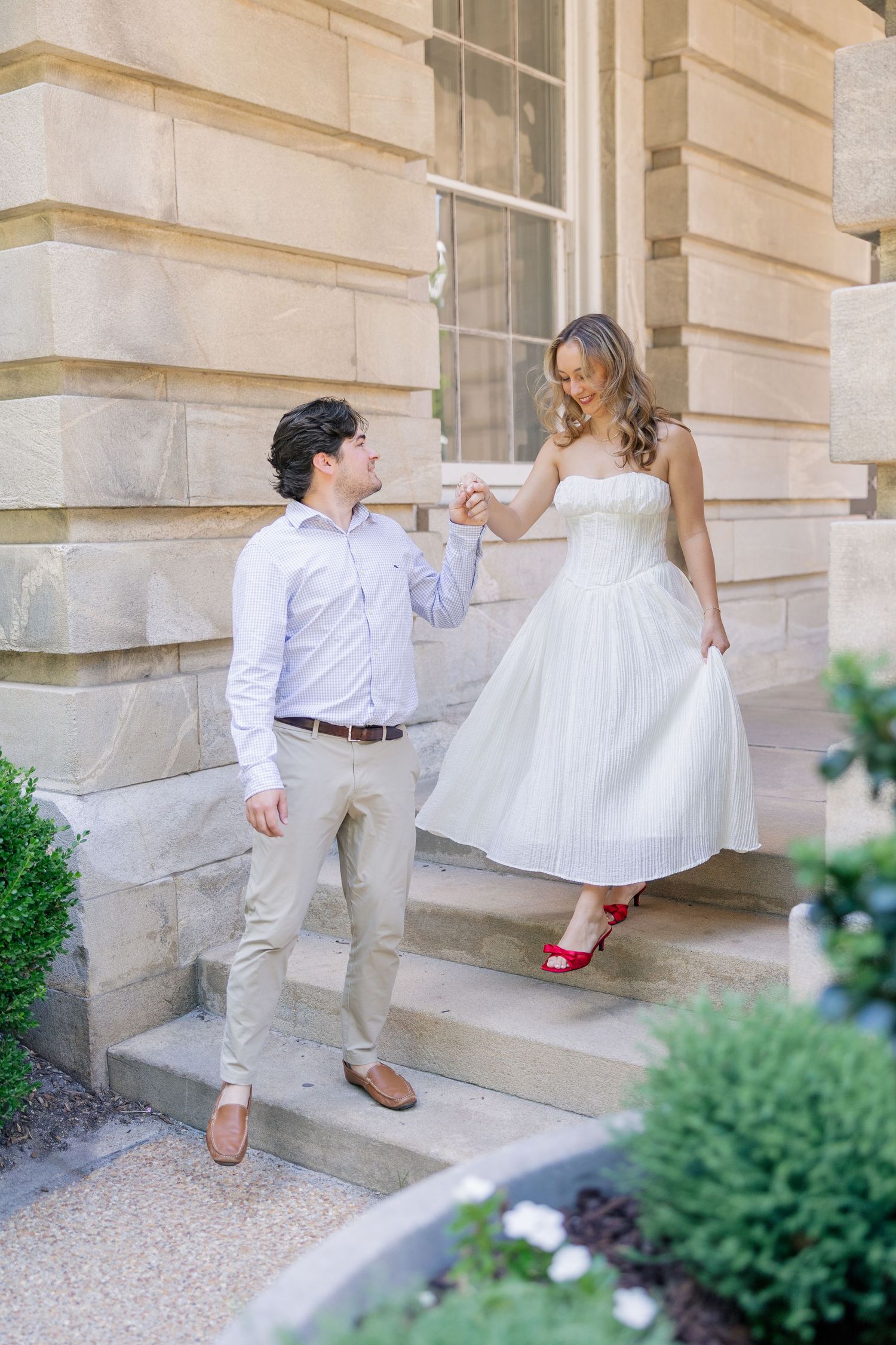 Couple holding hands on steps of historic stone building, woman wearing white dress and red heels.