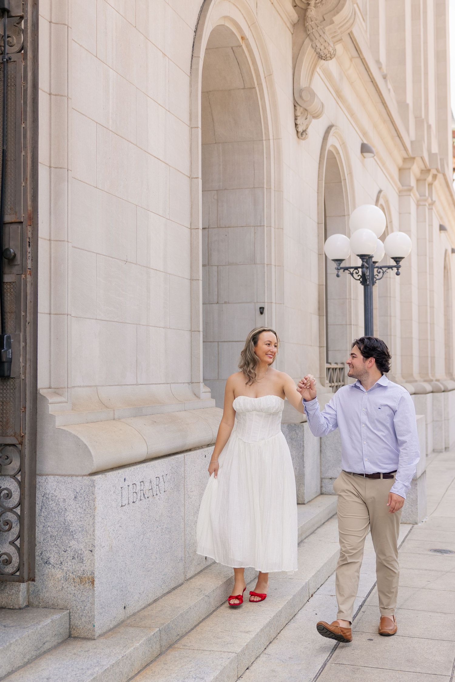 Couple in white dress and red heels walks hand-in-hand on library steps in elegant urban setting.
