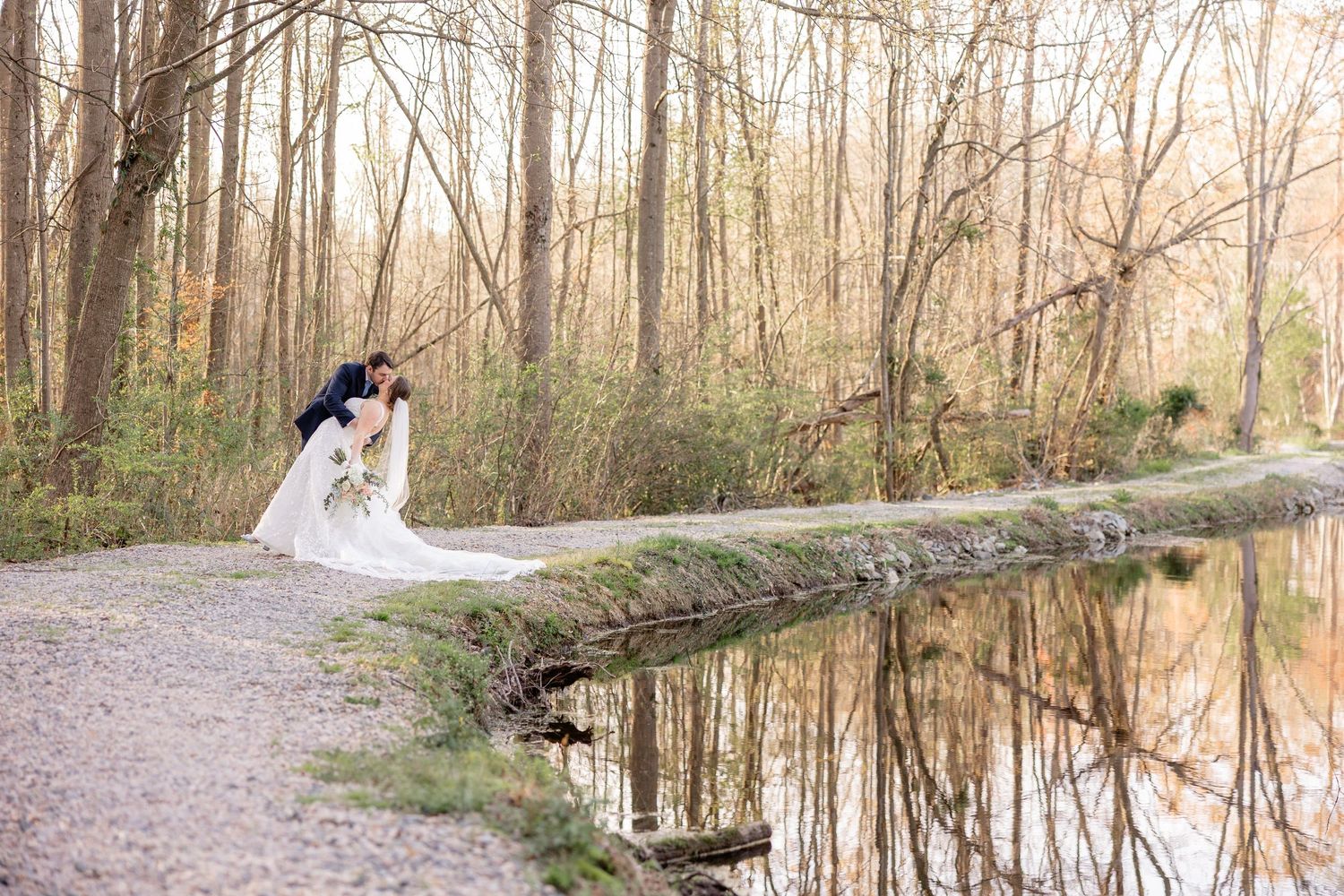 Someone in white bridal dress stands by a fallen log near a tranquil pond path in a peaceful wooded setting.