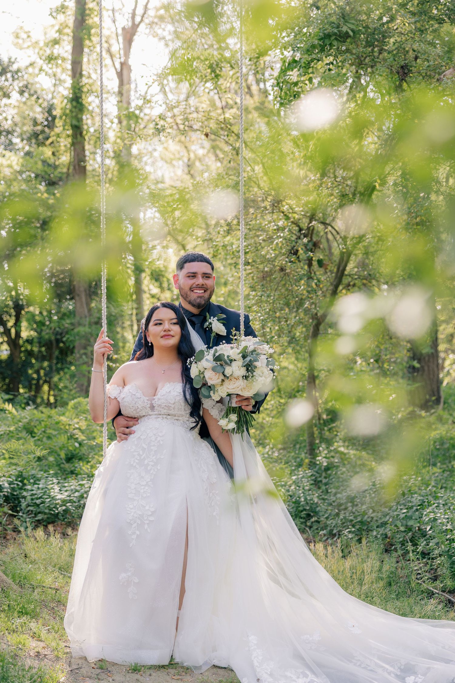 A joyful wedding couple poses with a bouquet in front of spring foliage and bokeh lighting effects.