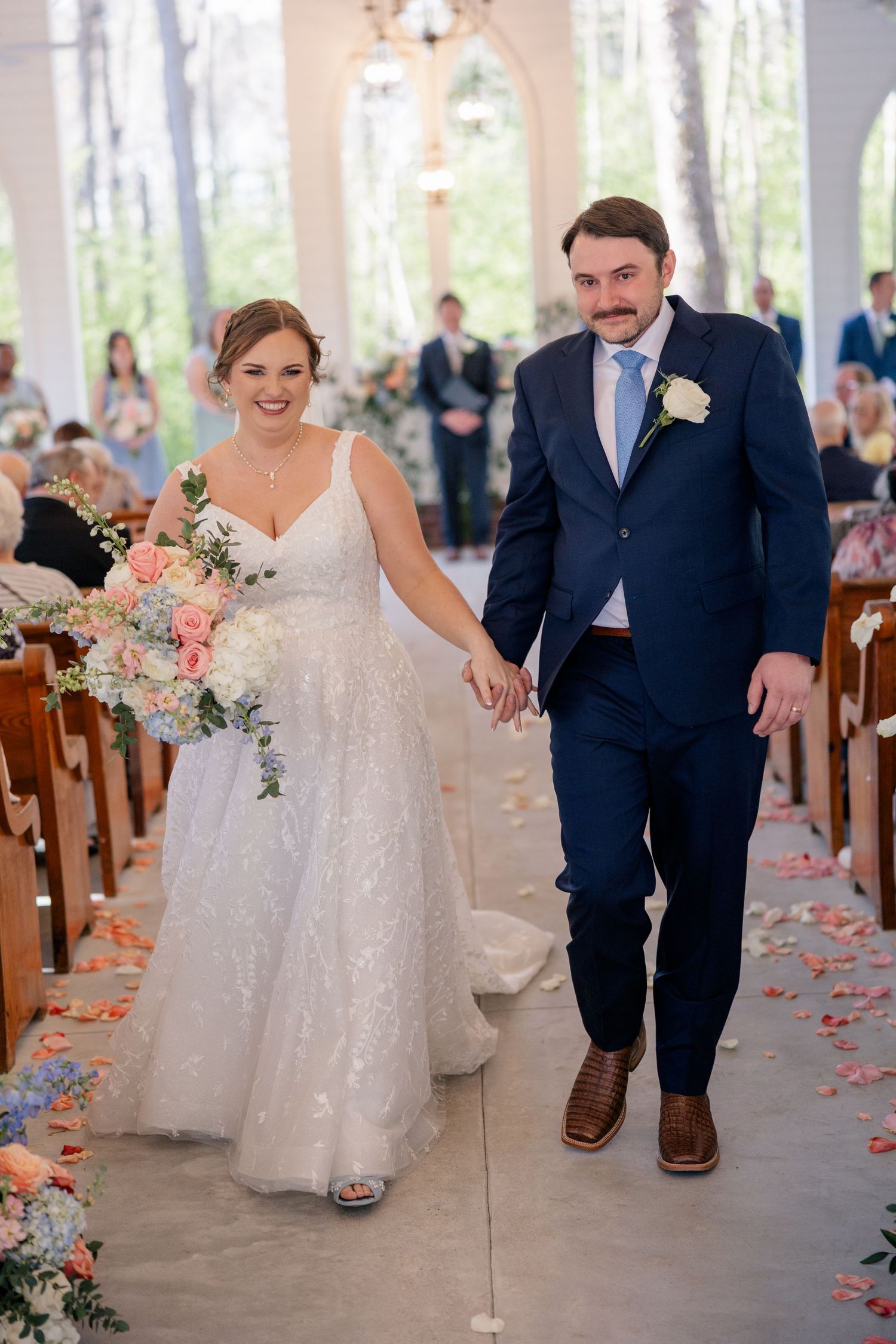 Newlyweds walk down the aisle together surrounded by floral arrangements and smiling guests.