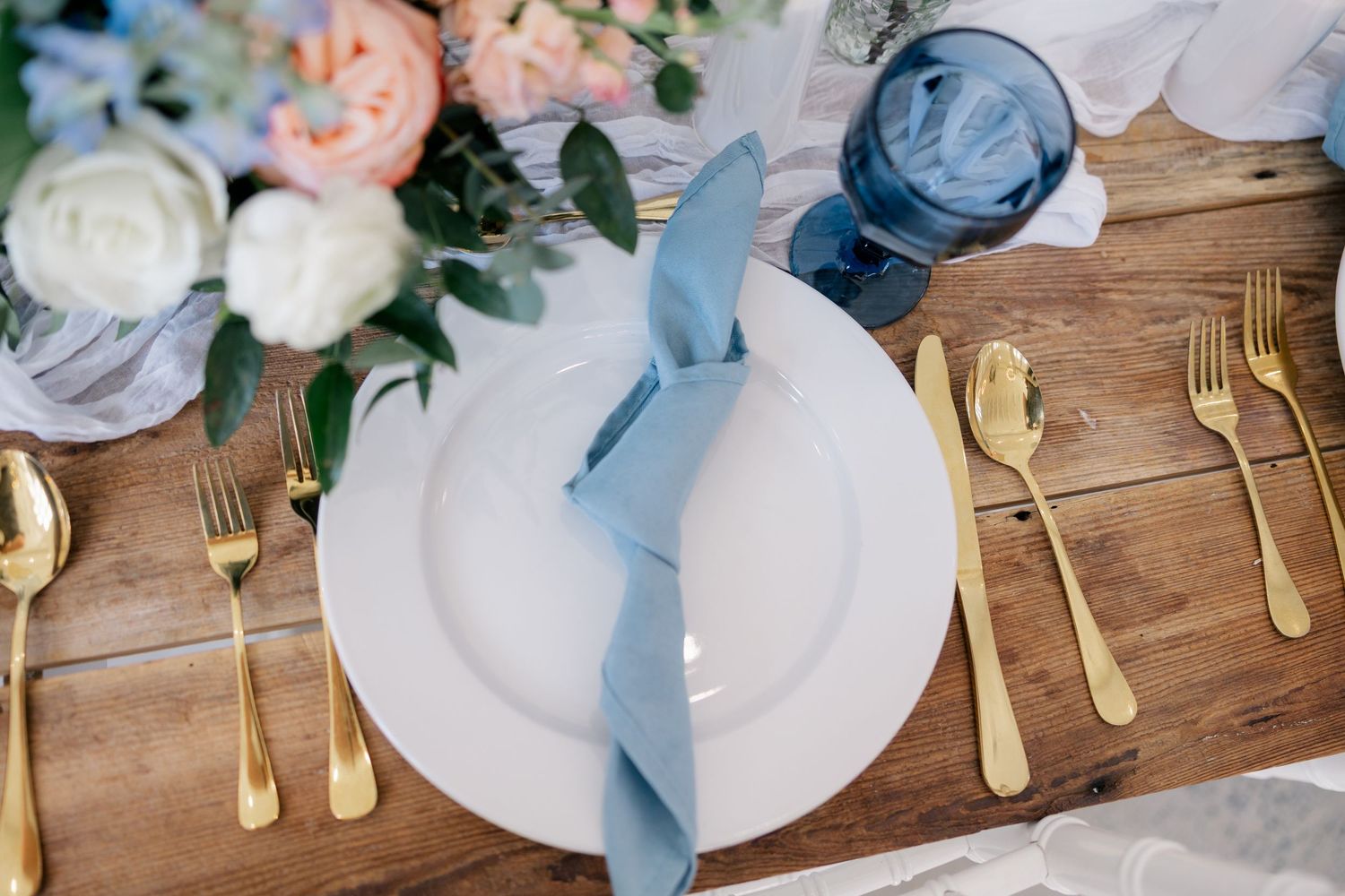 Place settings with white plates, gold utensils, and blue napkins on a rustic wooden table.