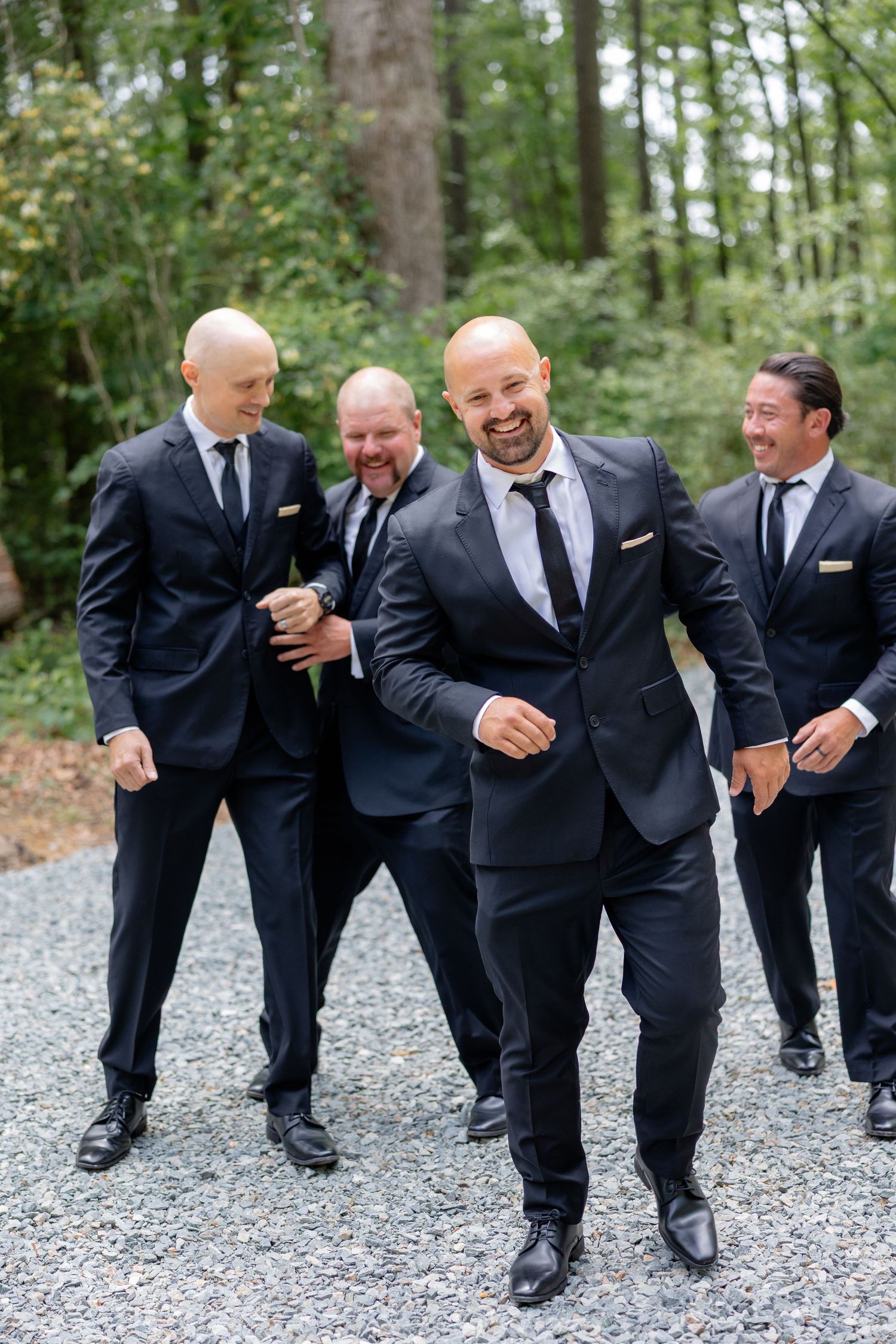 Wedding party members in dark suits share laughs while posing for casual photos on a gravel pathway.
