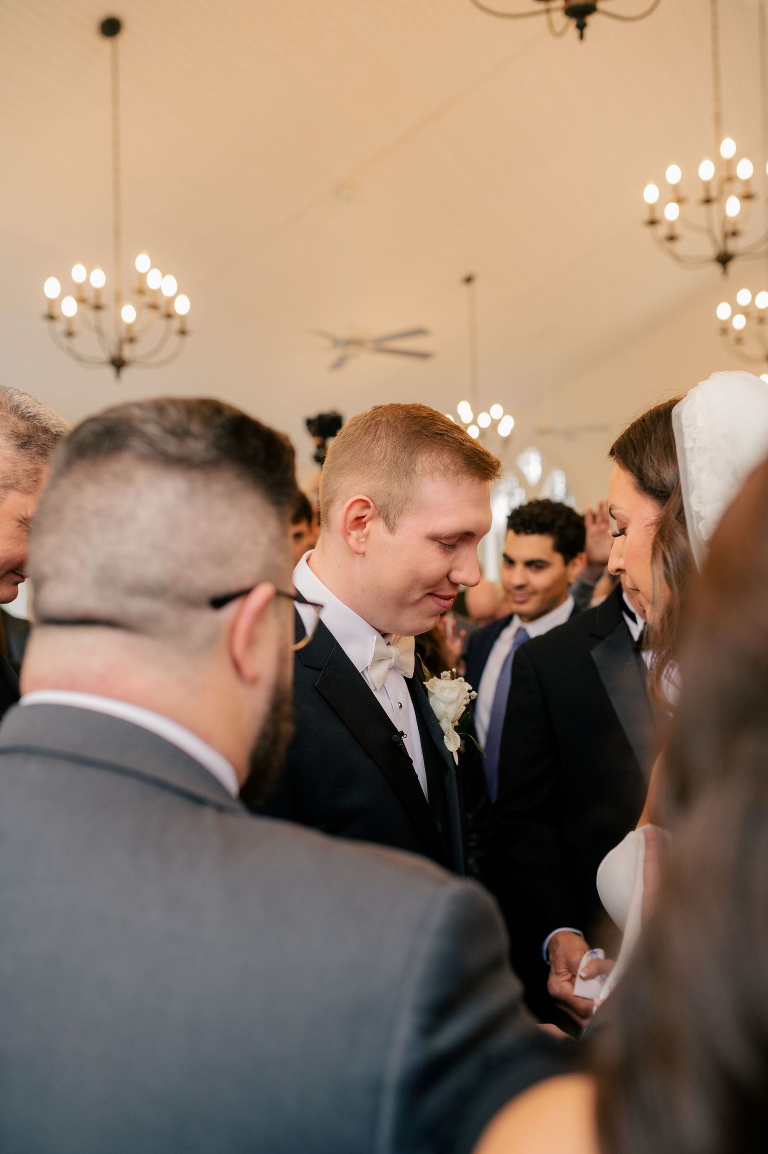 Wedding guests dressed in formal attire gathered in an elegant reception room with crystal chandeliers overhead.