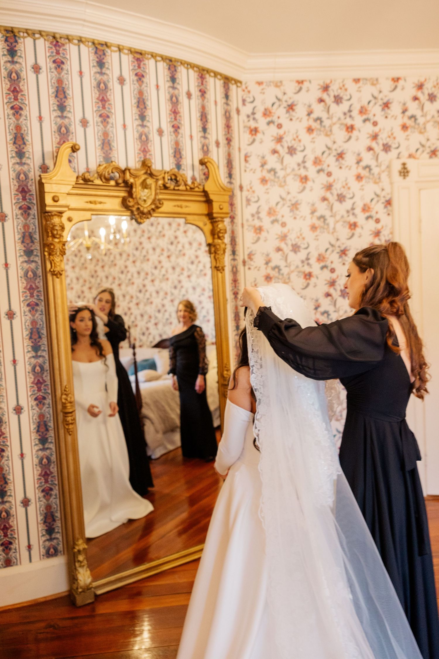 A wedding dress is being adjusted in an elegant room with floral wallpaper and gold-framed mirrors.