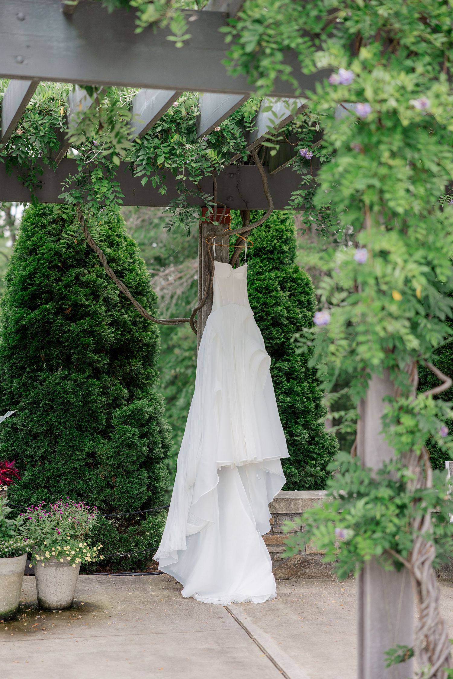 A white wedding dress hangs elegantly from a wooden pergola surrounded by lush greenery.