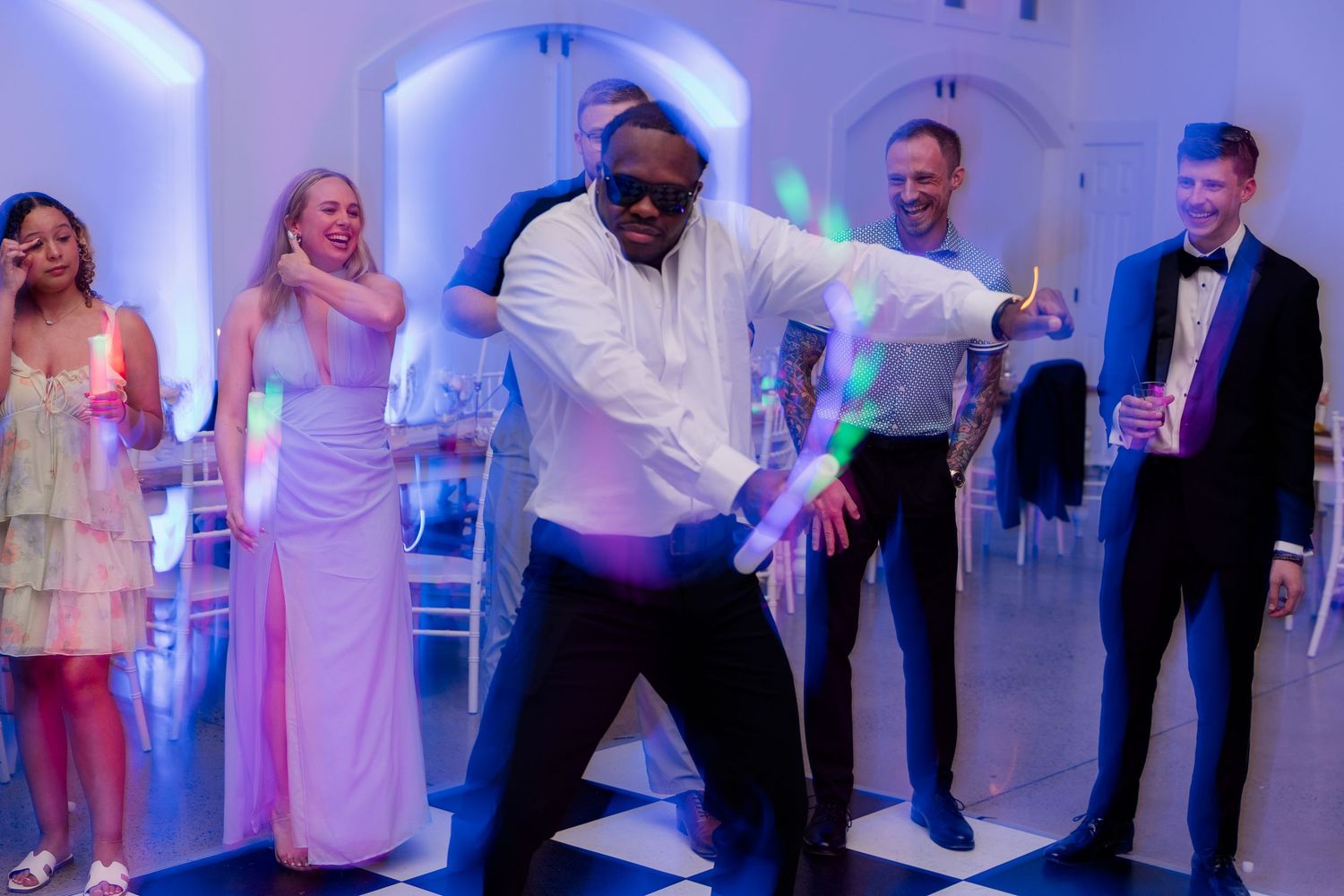 Wedding party dances in a line formation under blue accent lighting at indoor reception.