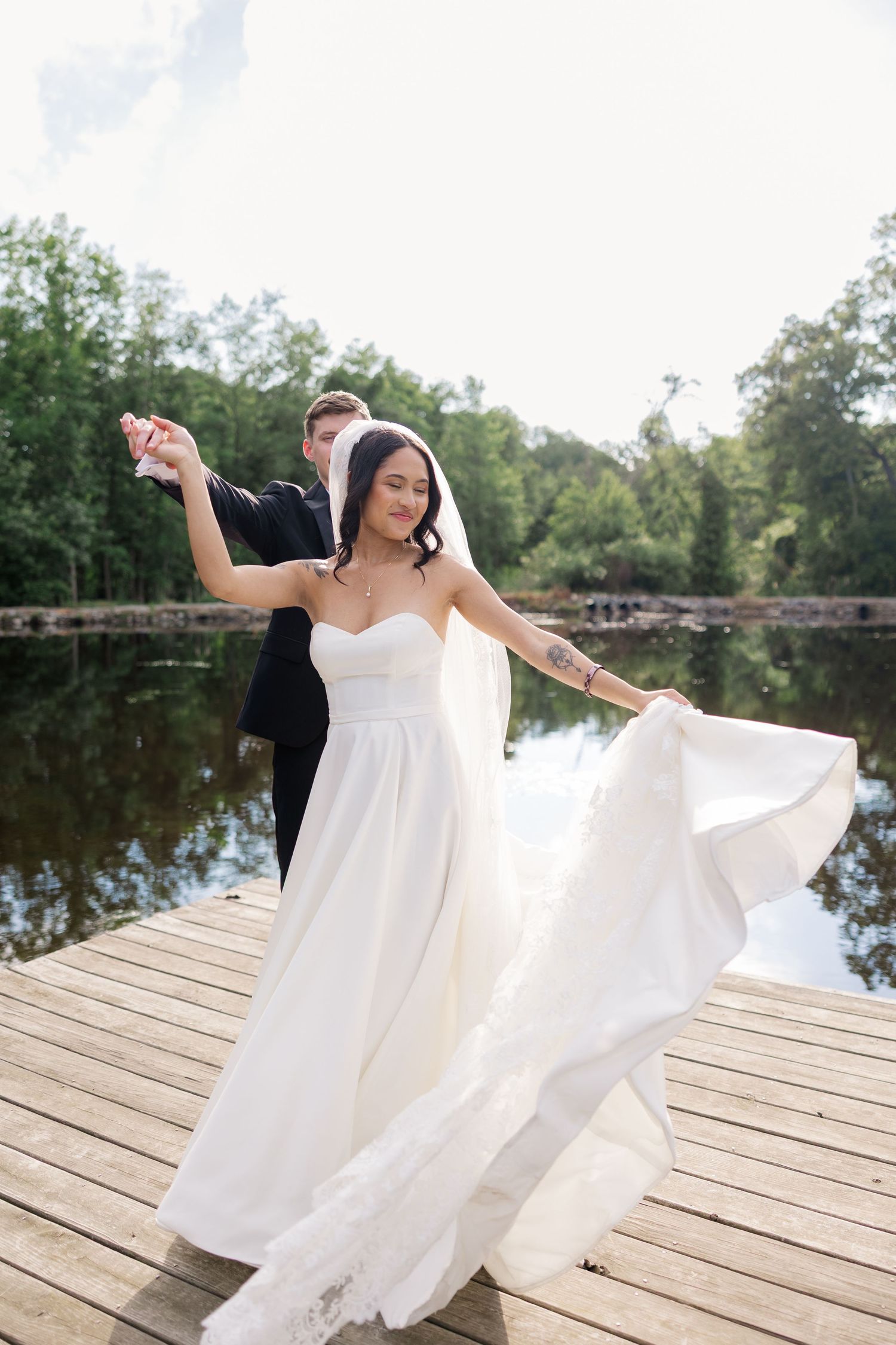 Bride in flowing white dress twirls gracefully on wooden dock by serene lake.