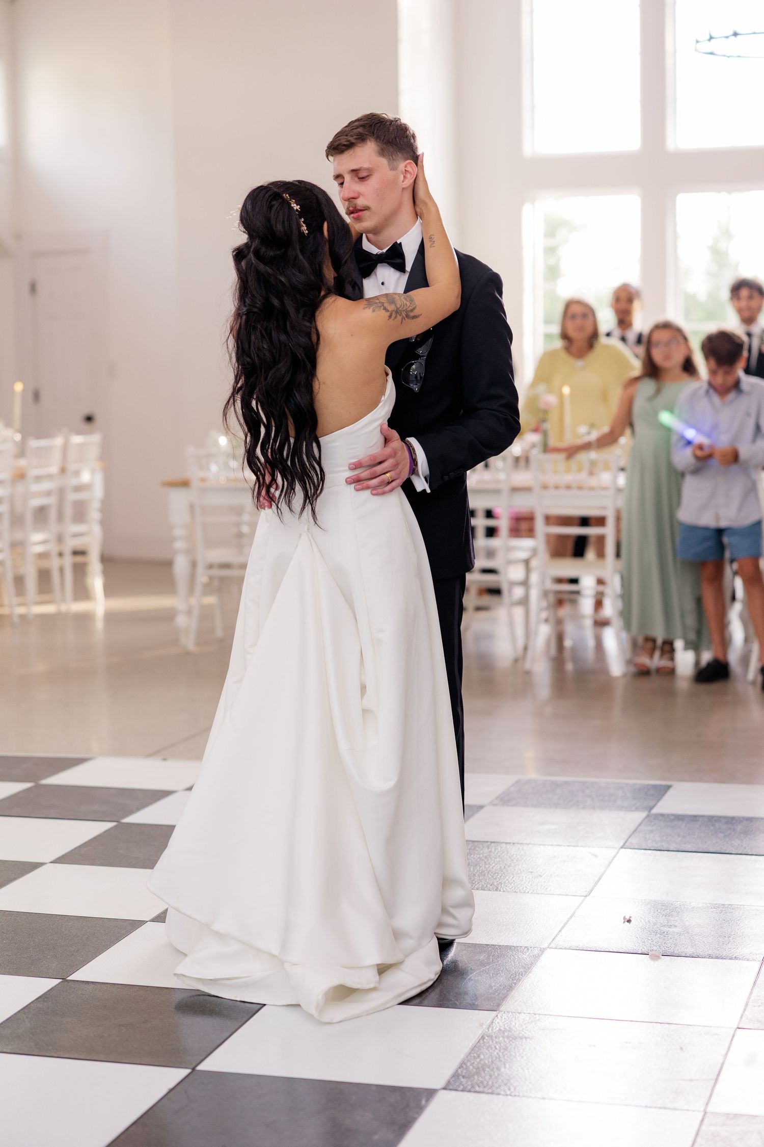 A romantic first dance between newlyweds on a black and white checkered floor with guests watching in background.