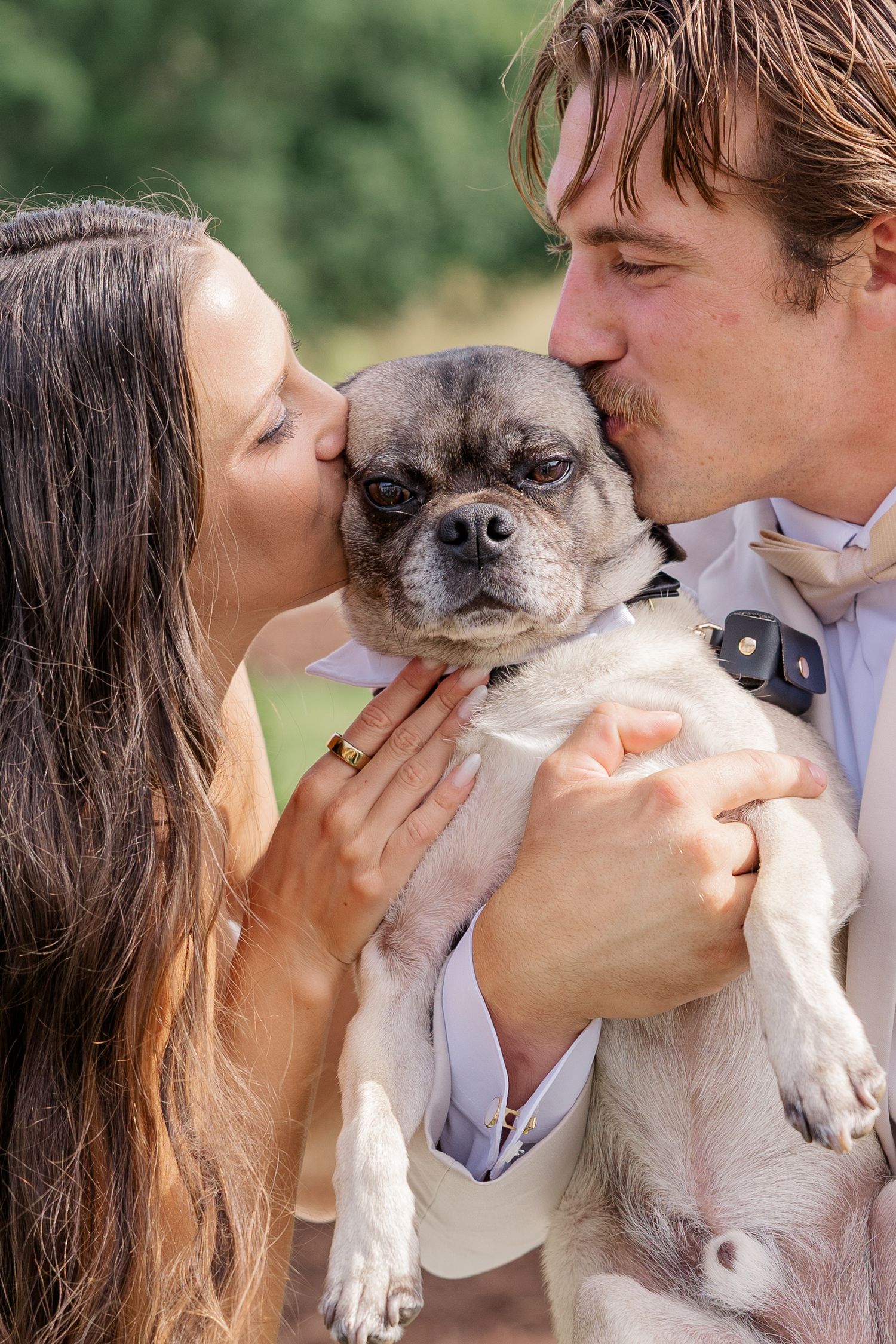 A couple kisses their small pug dog from both sides while dressed in formal attire.