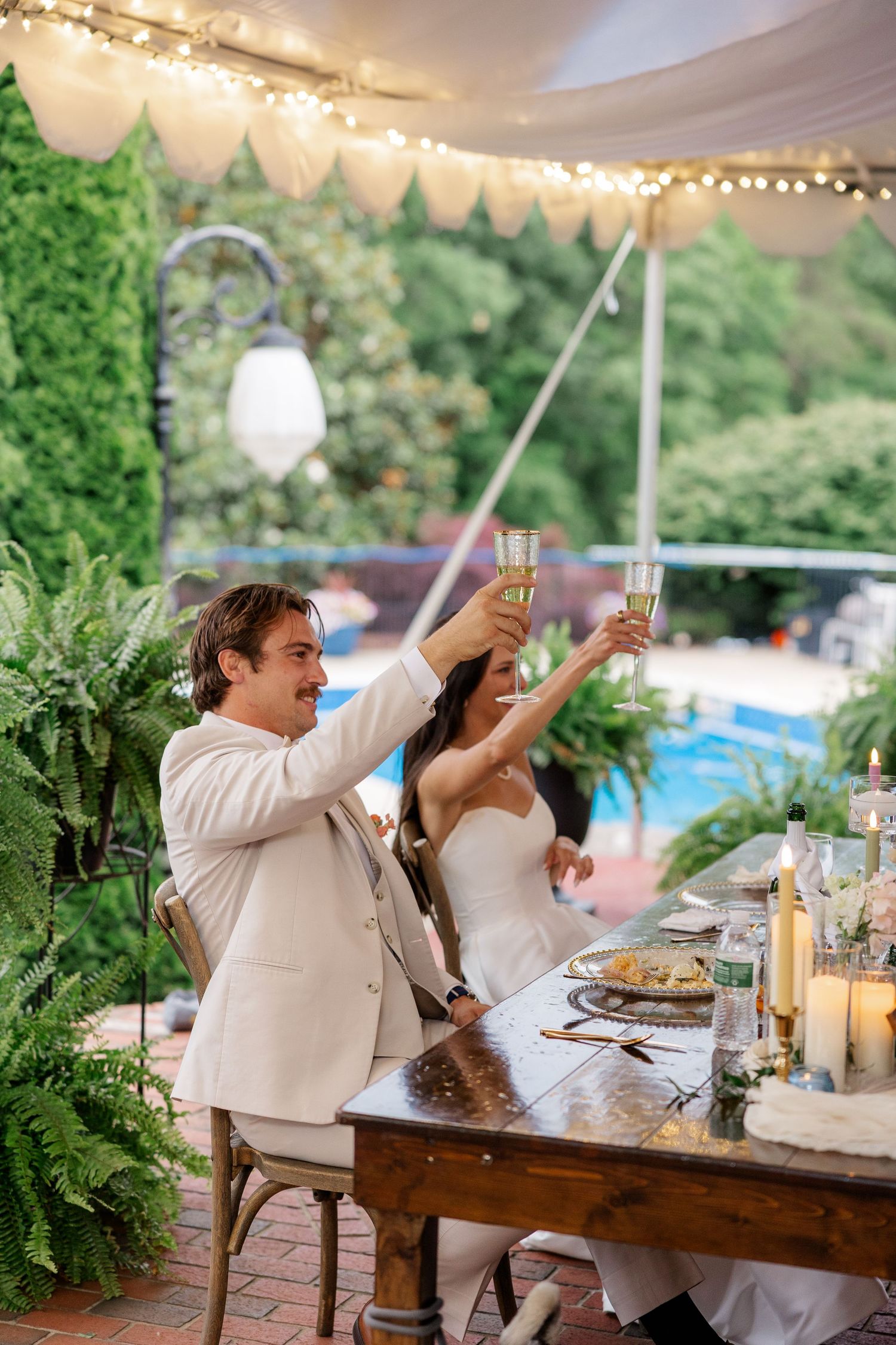 Newlyweds enjoy drinks and celebrate at a rustic wooden table under string lights at their reception.