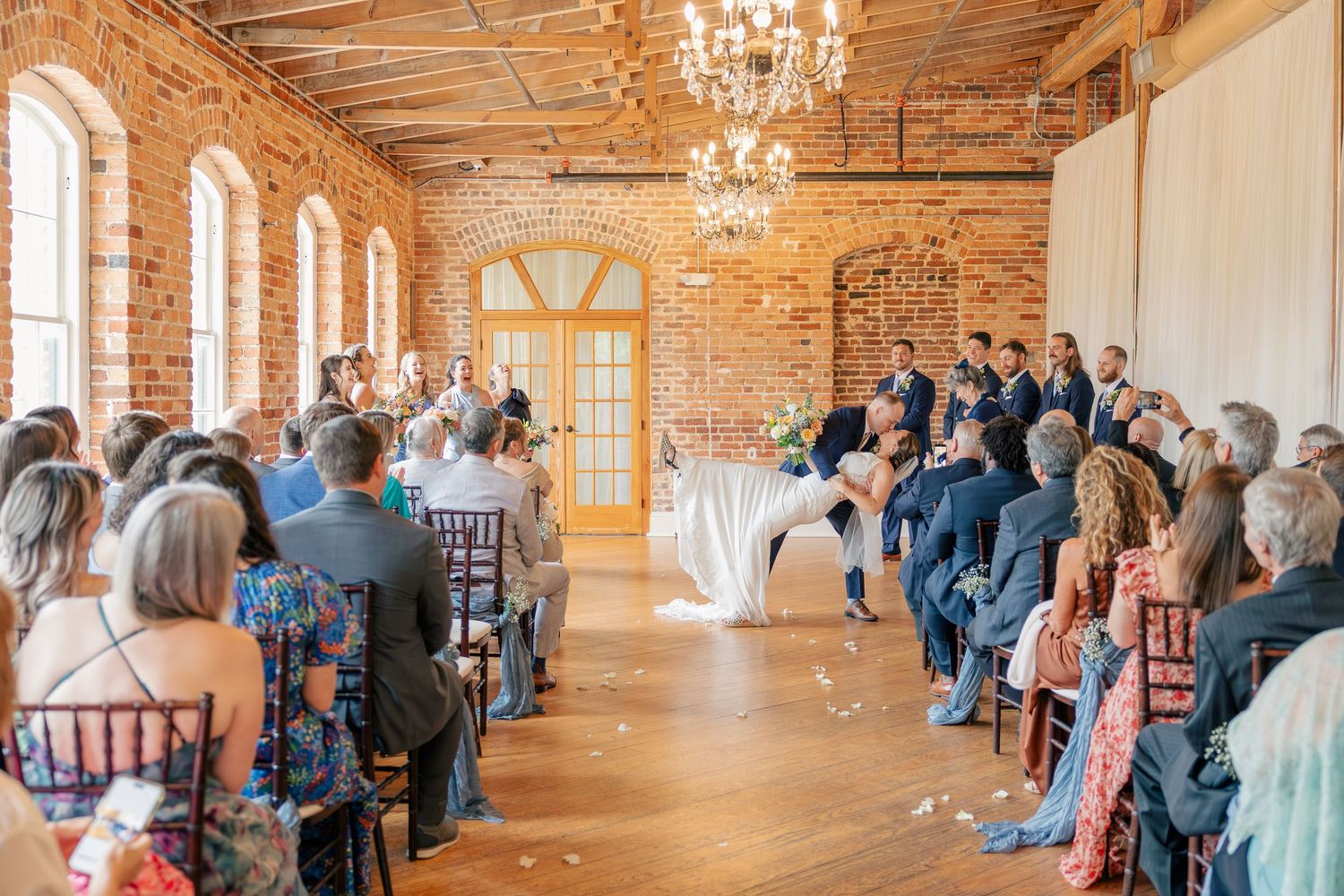 Wedding ceremony in a sunlit rustic brick venue with guests seated in chiavari chairs facing the altar.