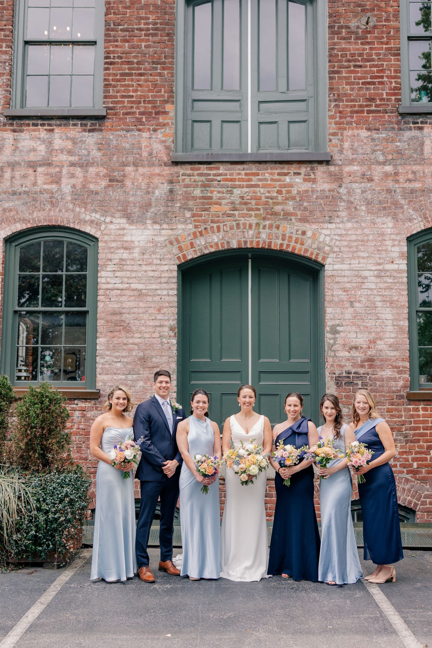 Bride and bridesmaids in blue gowns pose with groom before a rustic brick building with green doors.